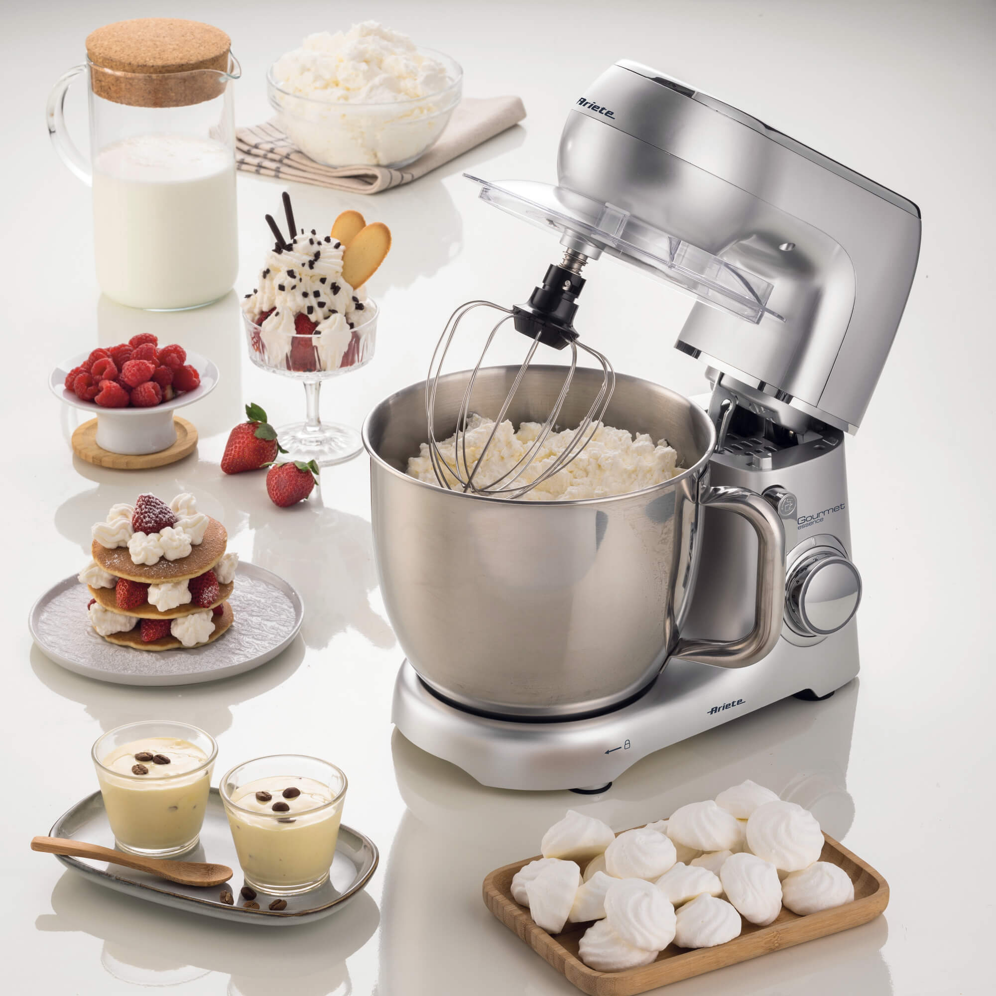 Ein silberner Küchenmixer mit einer Schüssel Sahne. Desserts wie Pfannkuchen, Himbeeren und Baiser sind im Bild.