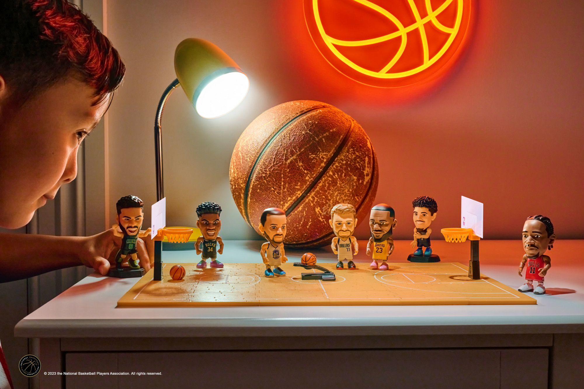 Ein Junge betrachtet Basketball-Wackelkopfpuppen auf einer Spielfläche. Im Hintergrund ein Basketball und ein Neon-Basketballschild.