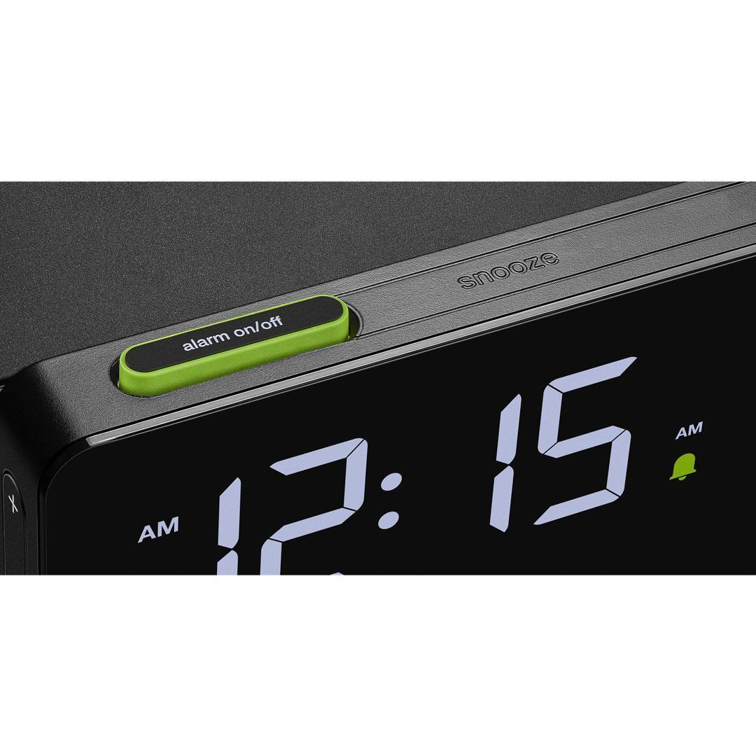 Eine Digitaluhr zeigt 12:15 Uhr morgens an. Ein grüner Knopf sagt Alarm ein/aus. Das Wort Schlummerfunktion ist sichtbar. Die Uhr ist schwarz.