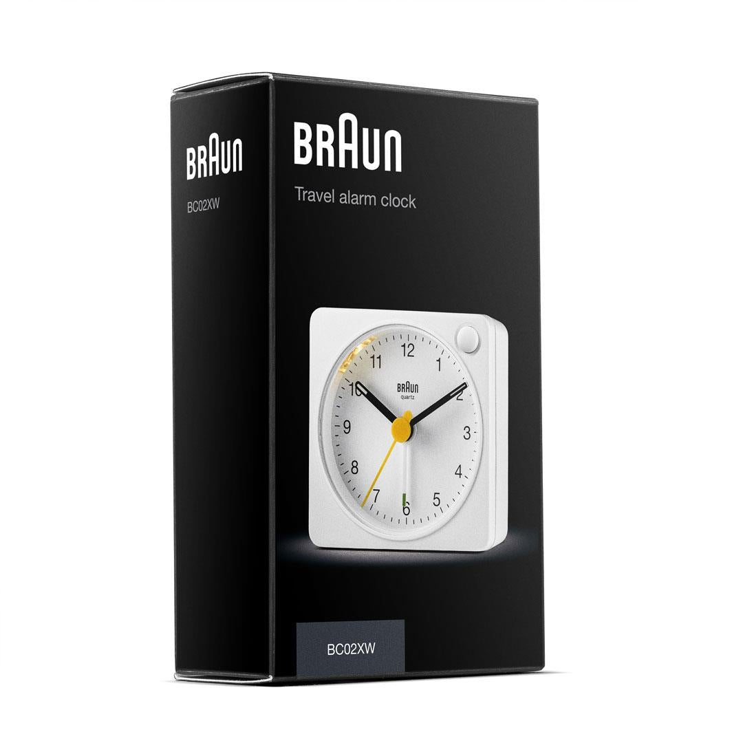 Una sveglia da viaggio Braun bianca nella sua scatola nera. Il quadrante ha una lancetta dei secondi gialla. La scatola ha il logo Braun.