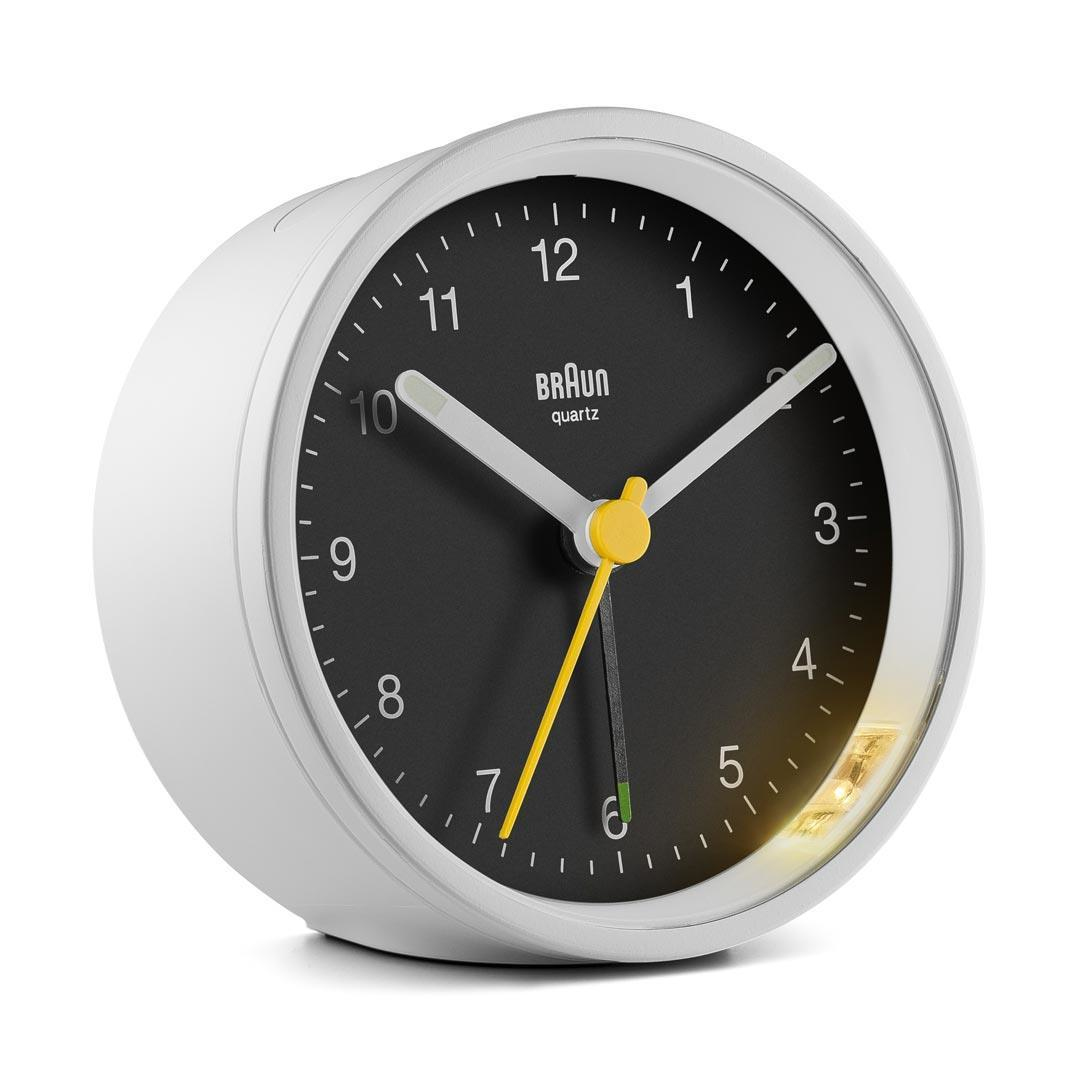Un réveil Braun quartz blanc avec cadran noir. Les aiguilles indiquent l'heure. Une aiguille et un bouton jaunes. Une lumière vive sur le côté.