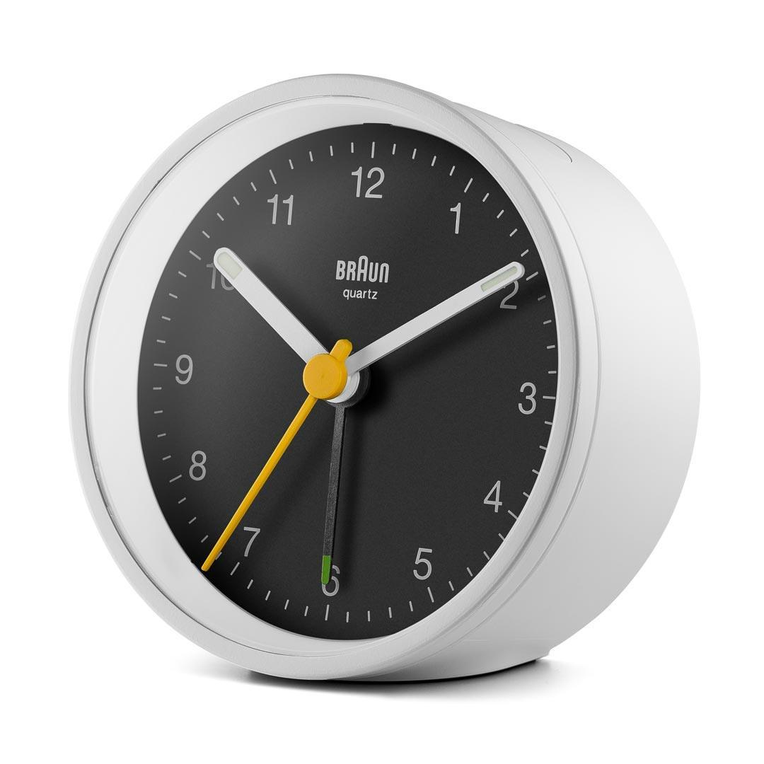 Un réveil Braun quartz blanc avec un cadran noir. Les aiguilles sont blanches et jaunes, indiquant 1:35. Les chiffres sont blancs.