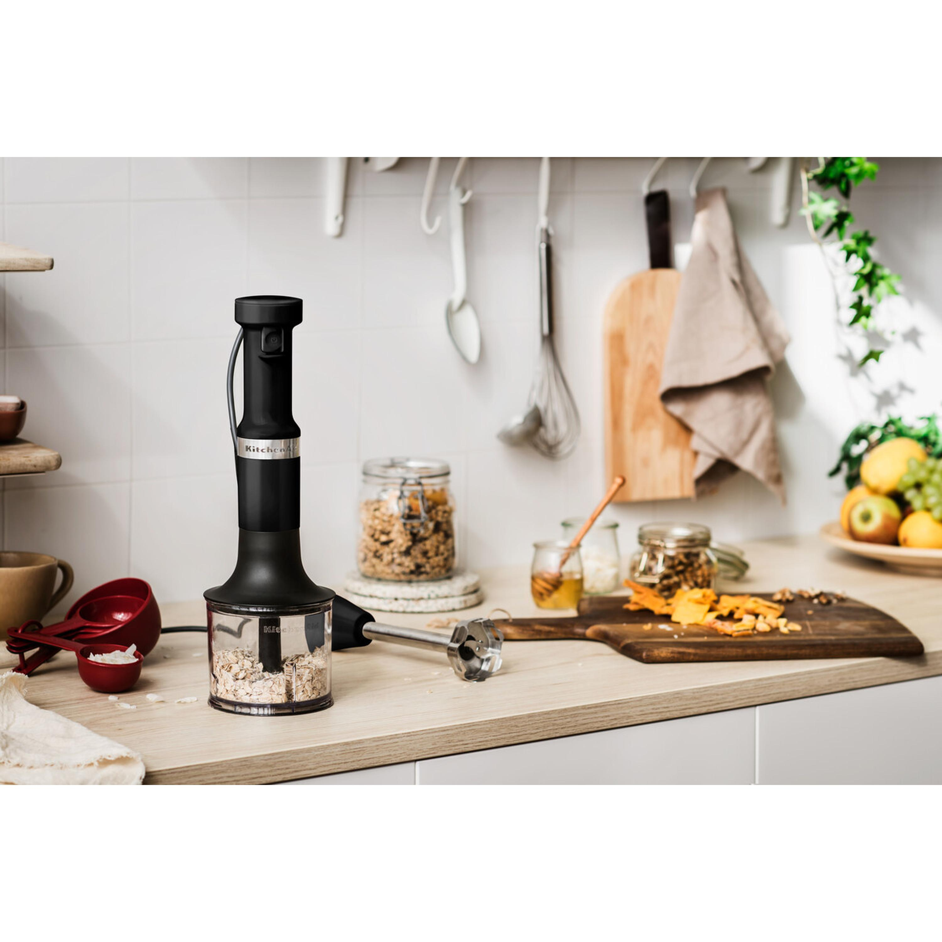 Black immersion blender on a countertop with ingredients. Background includes jars, cutting board, and fruit.