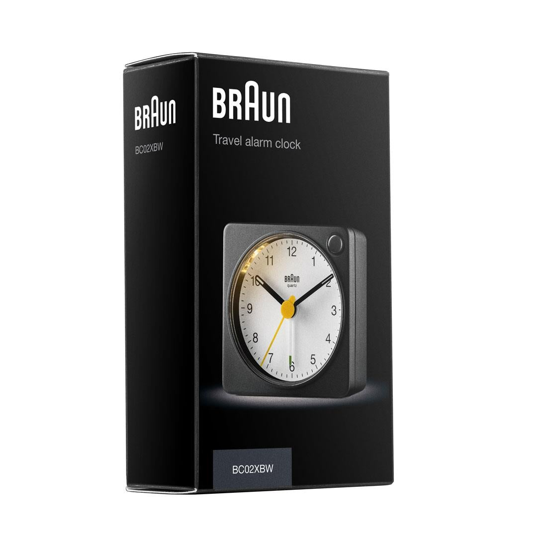 Un réveil de voyage Braun dans sa boîte. L'horloge est noire avec un cadran blanc et une trotteuse jaune. La boîte est noire.