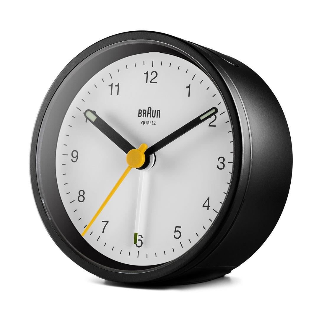 Un réveil Braun à quartz noir. Il a un cadran blanc avec des chiffres et des aiguilles noires. L'aiguille des secondes est jaune.