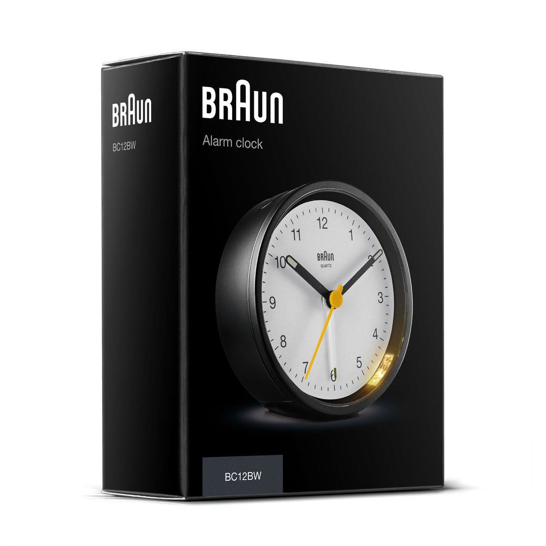 Un réveil Braun dans une boîte noire. Le cadran est blanc avec des chiffres noirs et des aiguilles jaunes. La boîte porte le logo de la marque.