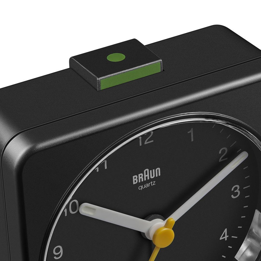 Primer plano de un despertador Braun Quartz negro. La esfera del reloj es negra con manecillas y números blancos. Hay un botón verde en la parte superior.