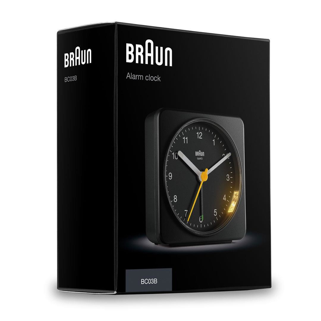 Un despertador Braun negro se muestra en su caja. La esfera del reloj es negra con números blancos y manecillas amarillas.