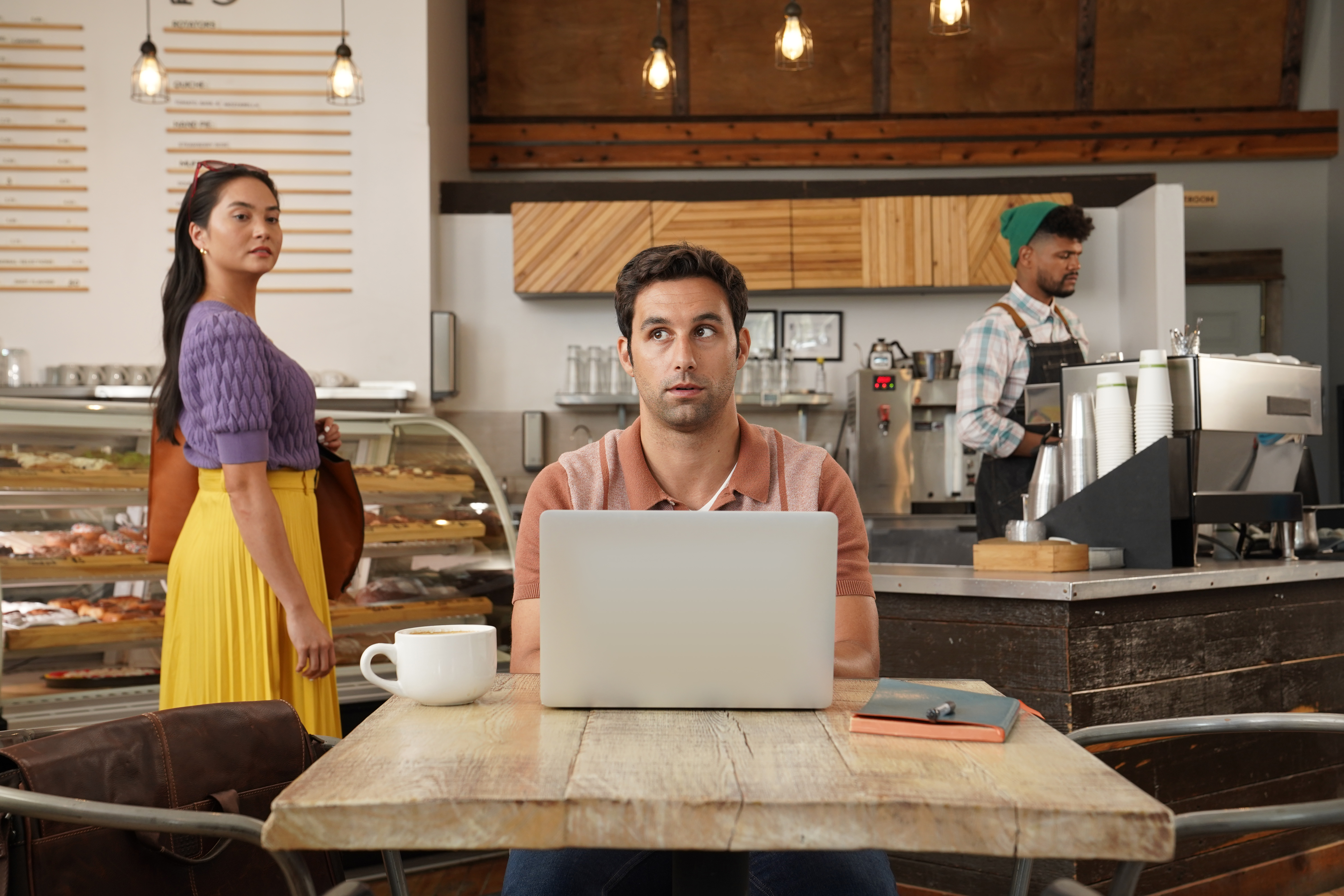 Ein Mann mit Laptop im Café blickt auf. Eine Frau steht bei Gebäck. Ein Barista arbeitet im Hintergrund.