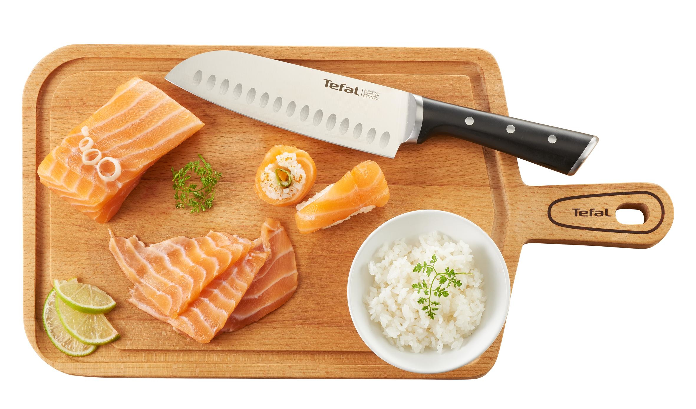 Ein Schneidebrett mit Lachs, Sushi, Reis und einem Messer. Limettenscheiben und Petersilie sind ebenfalls vorhanden. Das Messer ist mit Tefal beschriftet.