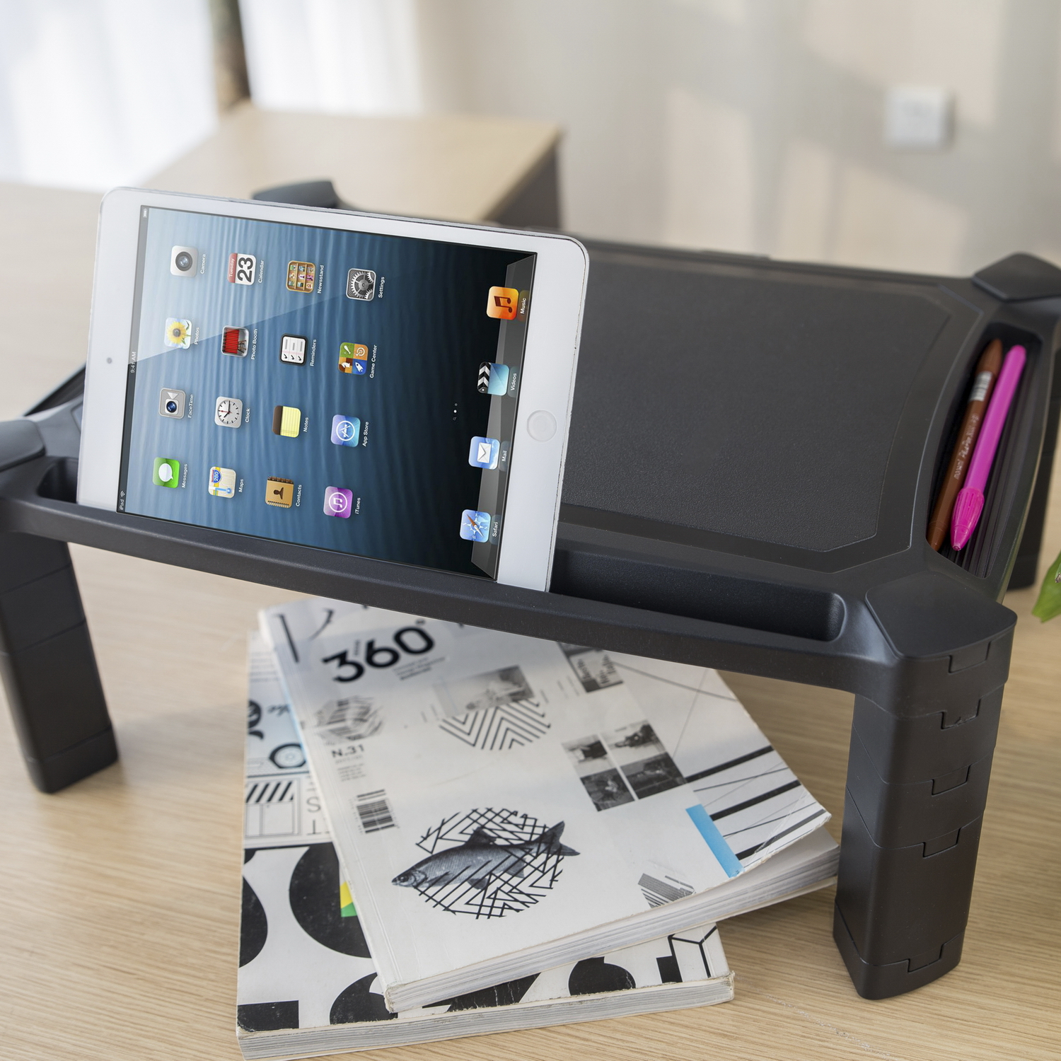 Un iPad sobre un soporte negro sobre revistas. Hay bolígrafos en una ranura. El iPad muestra iconos de aplicaciones. El fondo es un escritorio y una pared blanca.