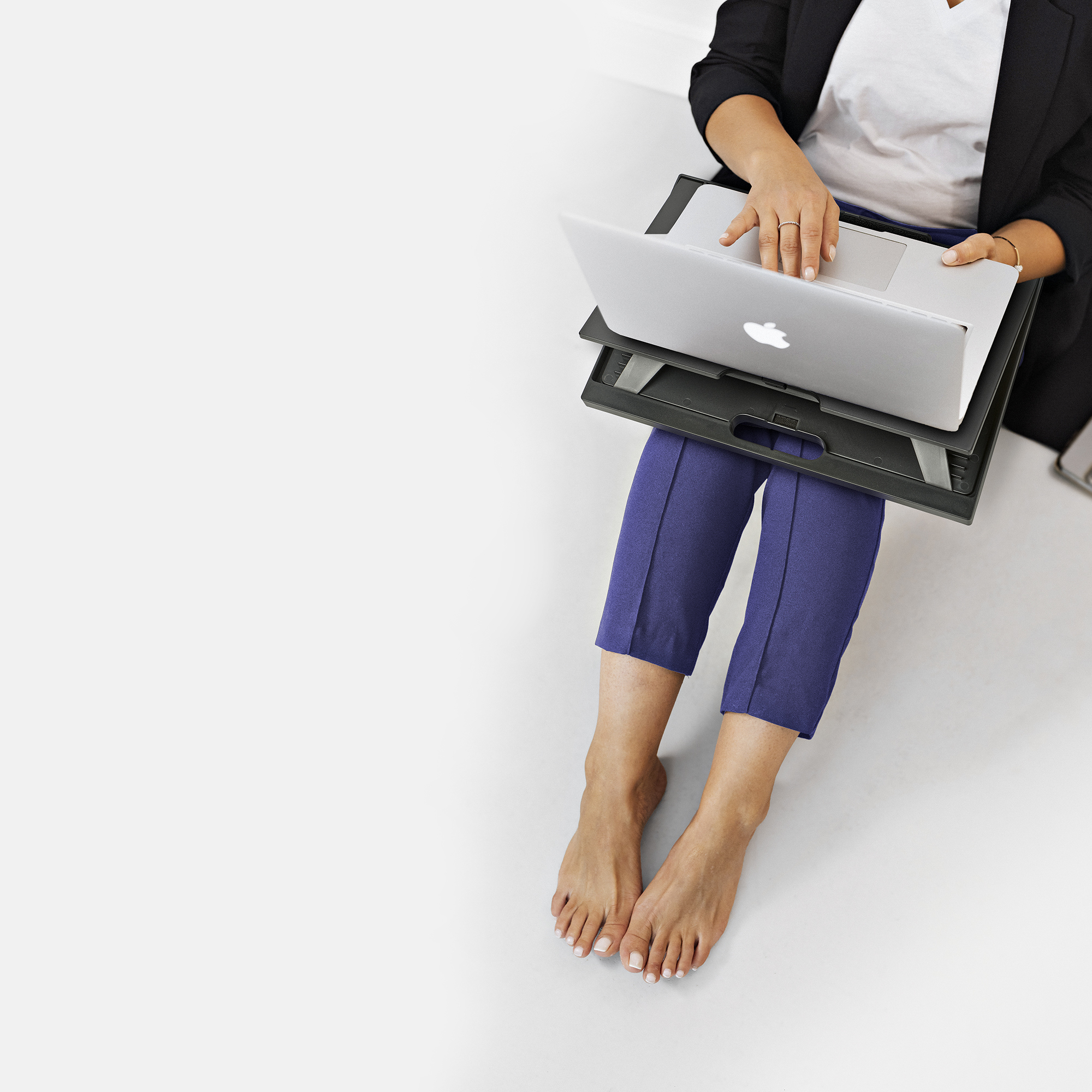 Person sitzt mit Laptop auf dem Schoß. Blaue Hose, schwarzer Blazer und weißes Hemd. Barfuß auf weißer Oberfläche.