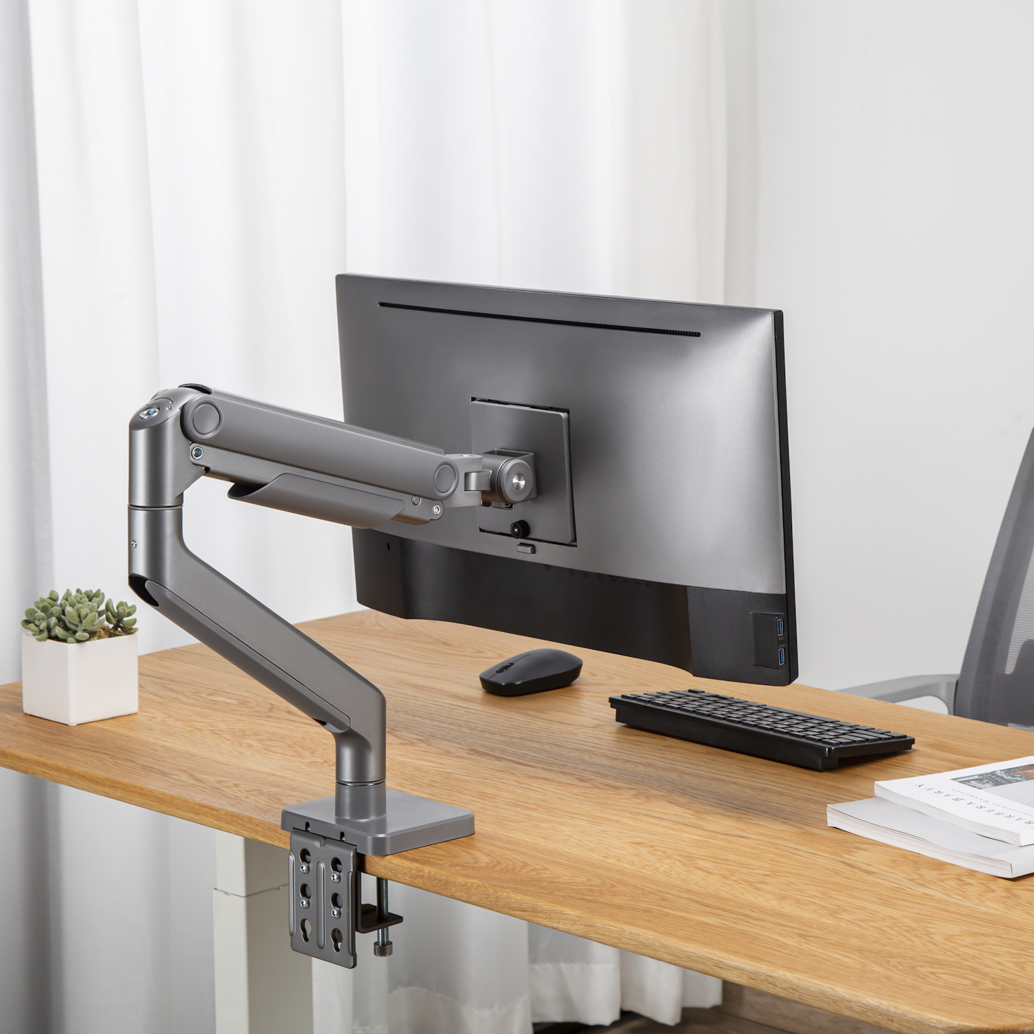 Un monitor per computer è montato su un braccio grigio su una scrivania di legno. Sulla scrivania ci sono anche una tastiera, un mouse e una pianta.