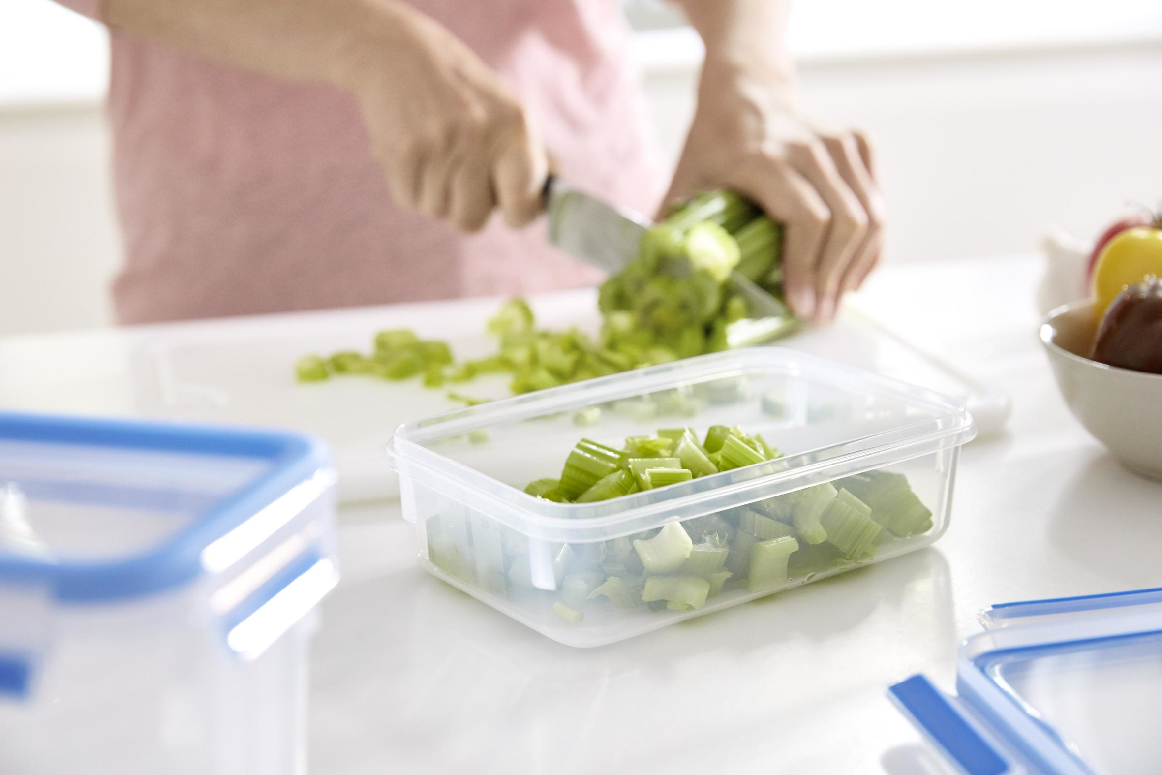 Osoba kroi seler na desce do krojenia. Posiekany seler znajduje się w pojemniku. Kolejny pojemnik jest na pierwszym planie.