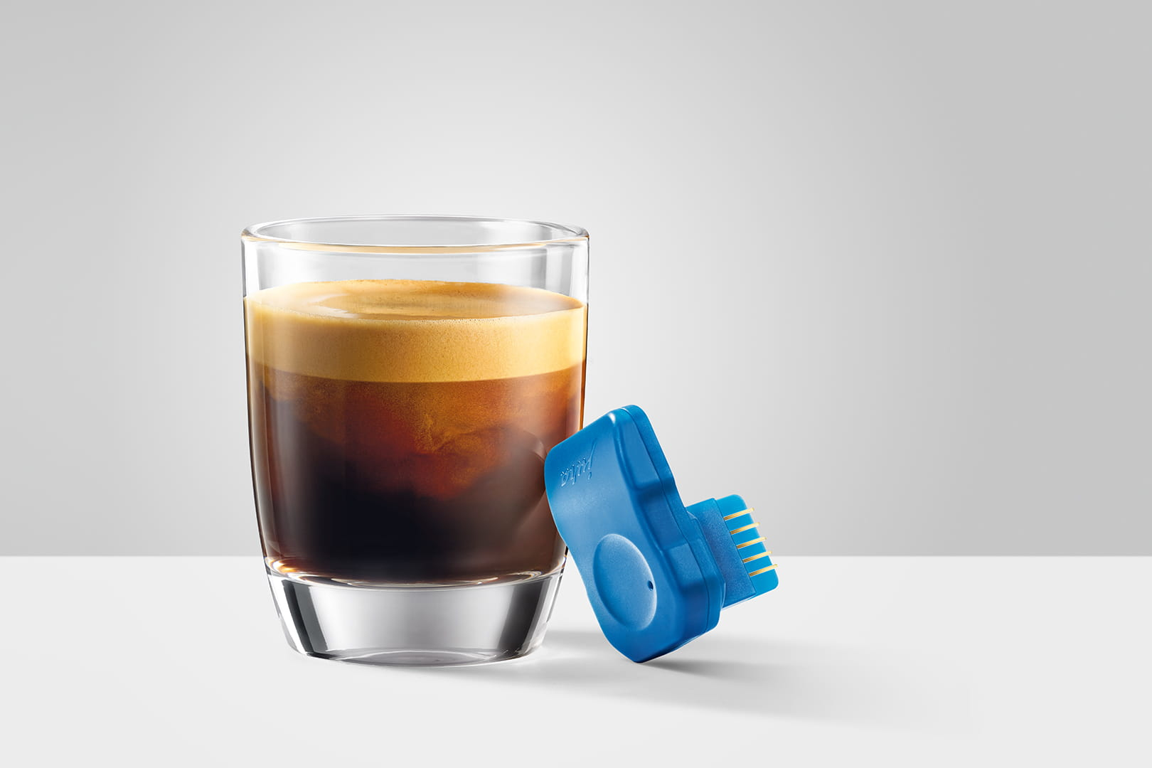 Ein Glas Espresso mit Crema, daneben ein blaues Gerät mit Goldpins auf weißer Oberfläche.
