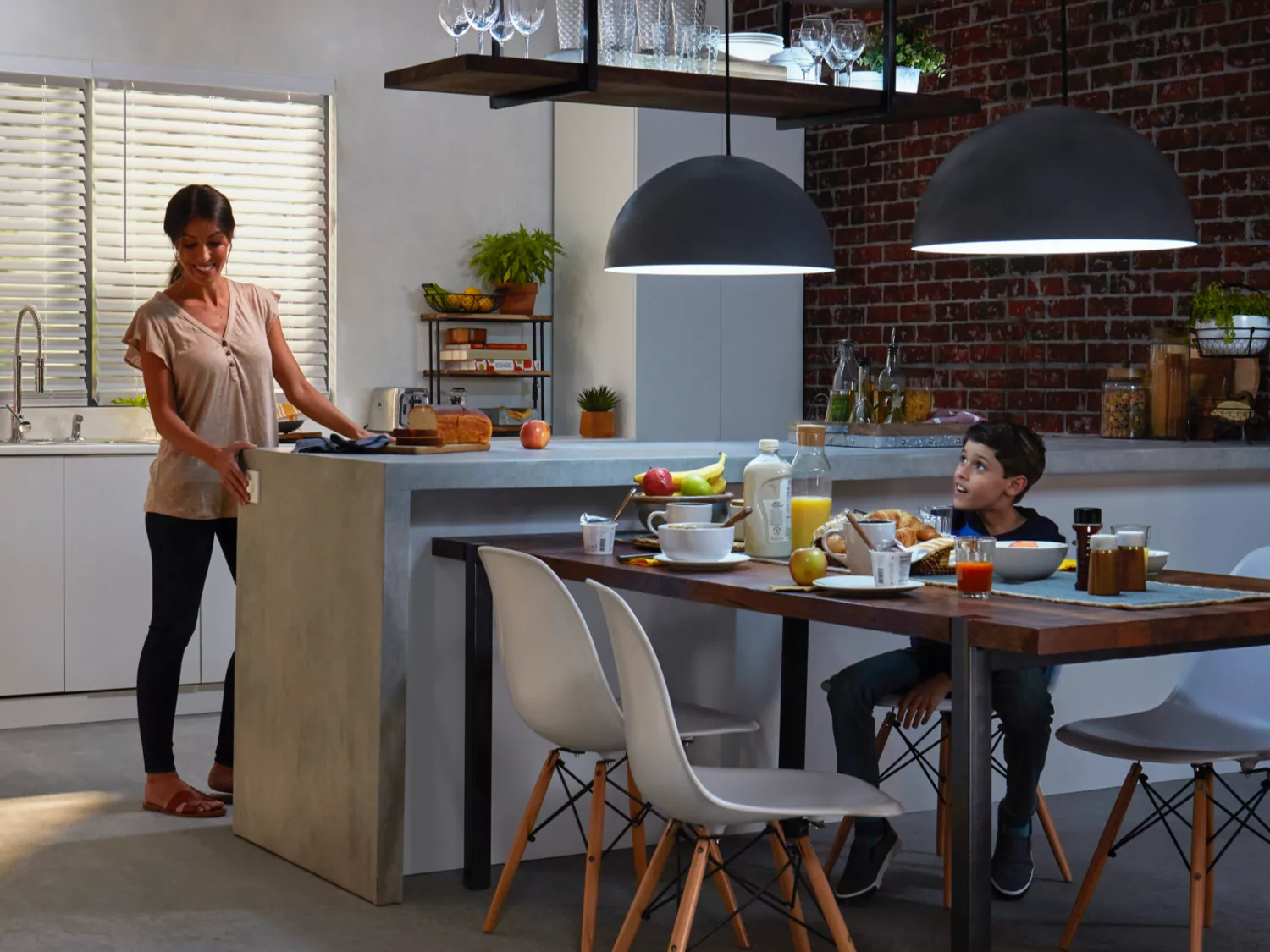 Kobieta i chłopiec w kuchni. Kobieta jest blisko blatu. Chłopiec siedzi przy stole z śniadaniem.