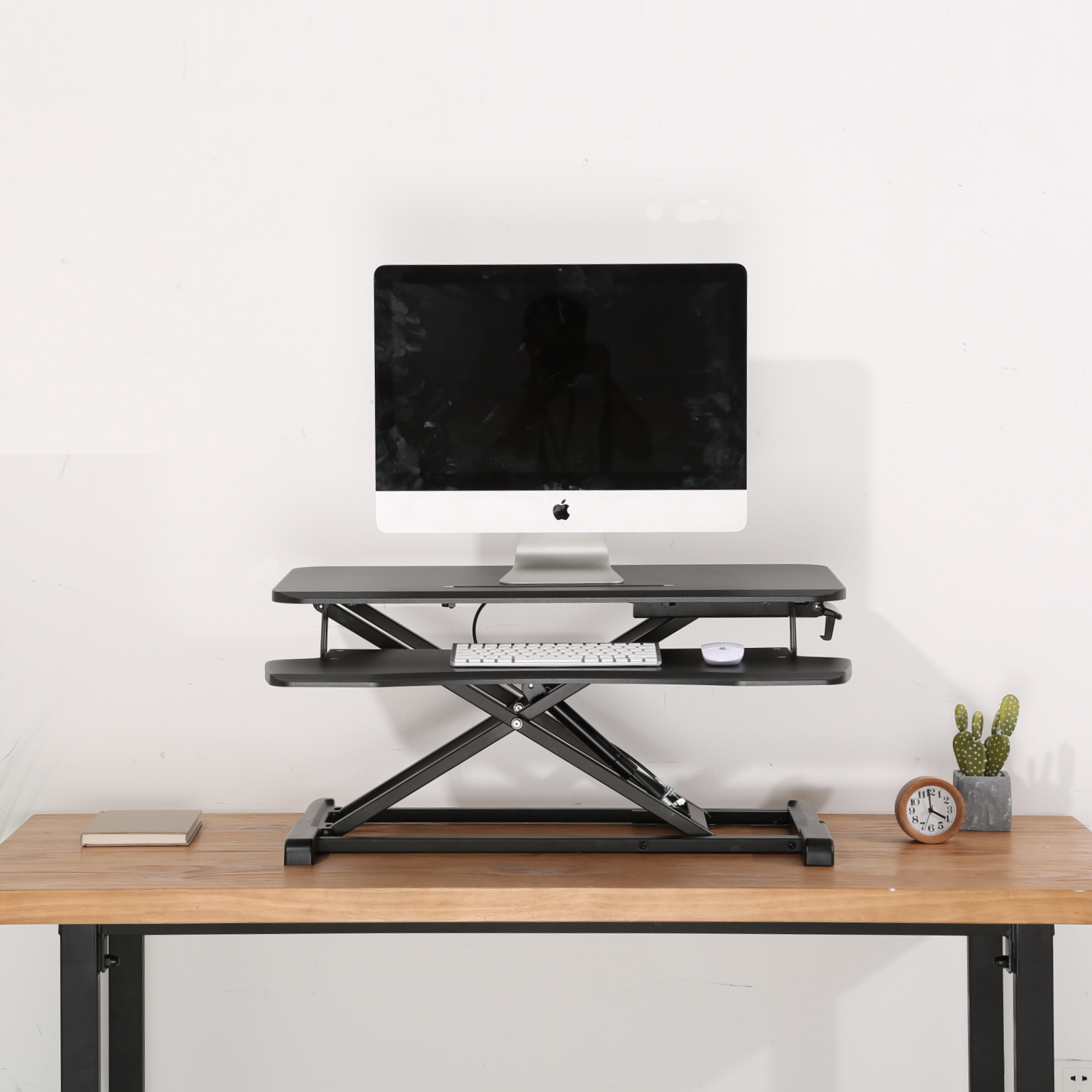 Una configuración de computadora en un escritorio. Un monitor se encuentra sobre un escritorio negro ajustable con teclado y ratón. Un reloj y un cactus están en el escritorio.