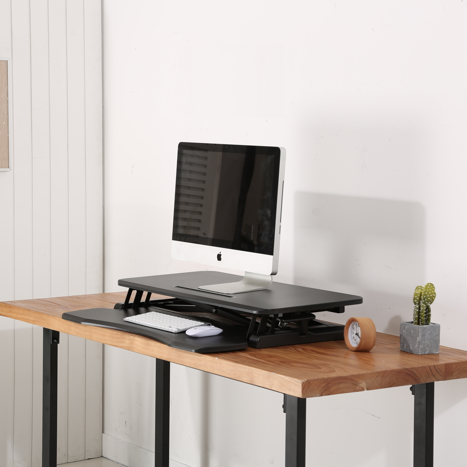 Una configuración de computadora en un escritorio de madera. Incluye un monitor, teclado, ratón, un reloj y un cactus. Pared blanca al fondo.