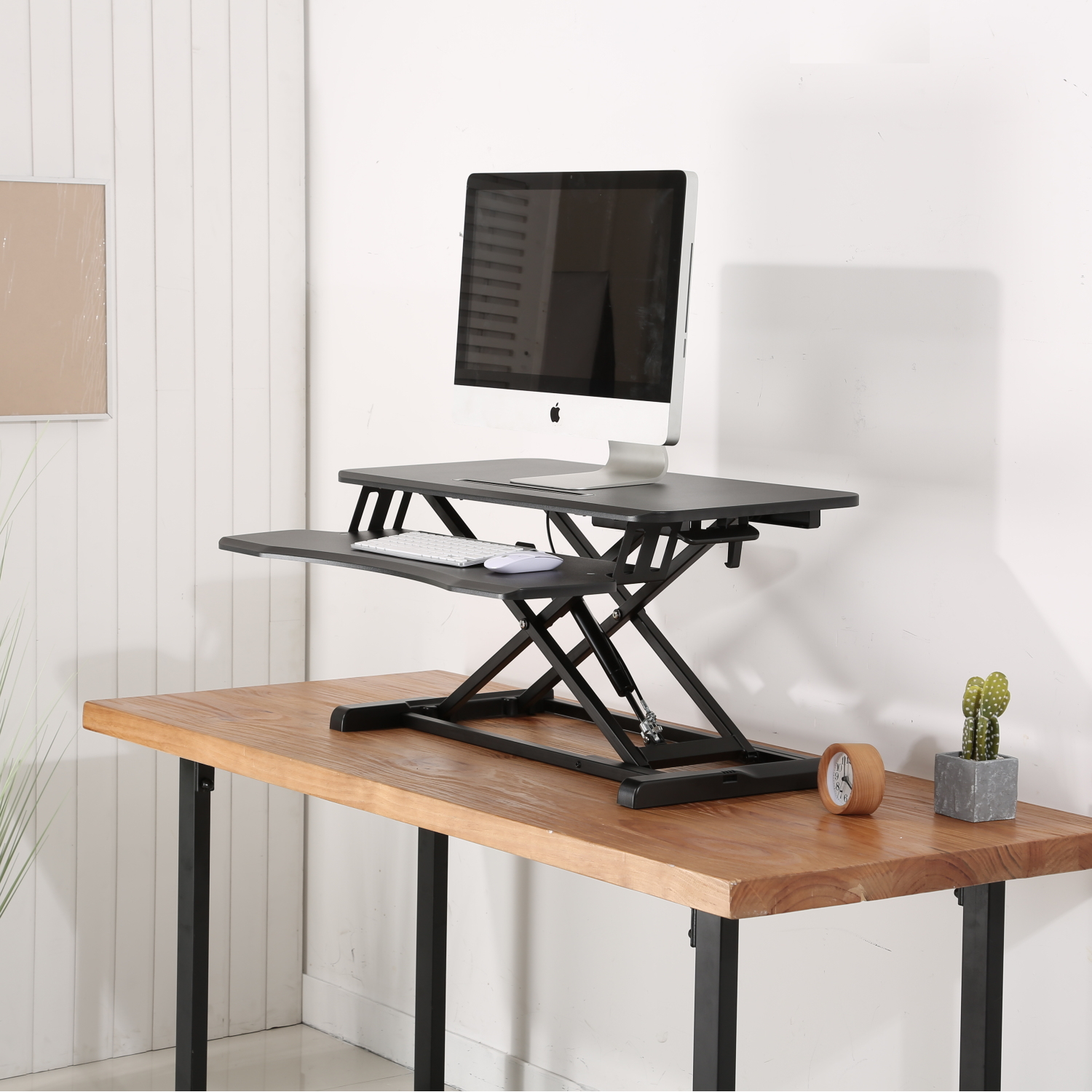 Una configuración de escritorio con computadora, escritorio de pie, teclado, ratón, reloj y cactus sobre una mesa de madera. Pared blanca al fondo.