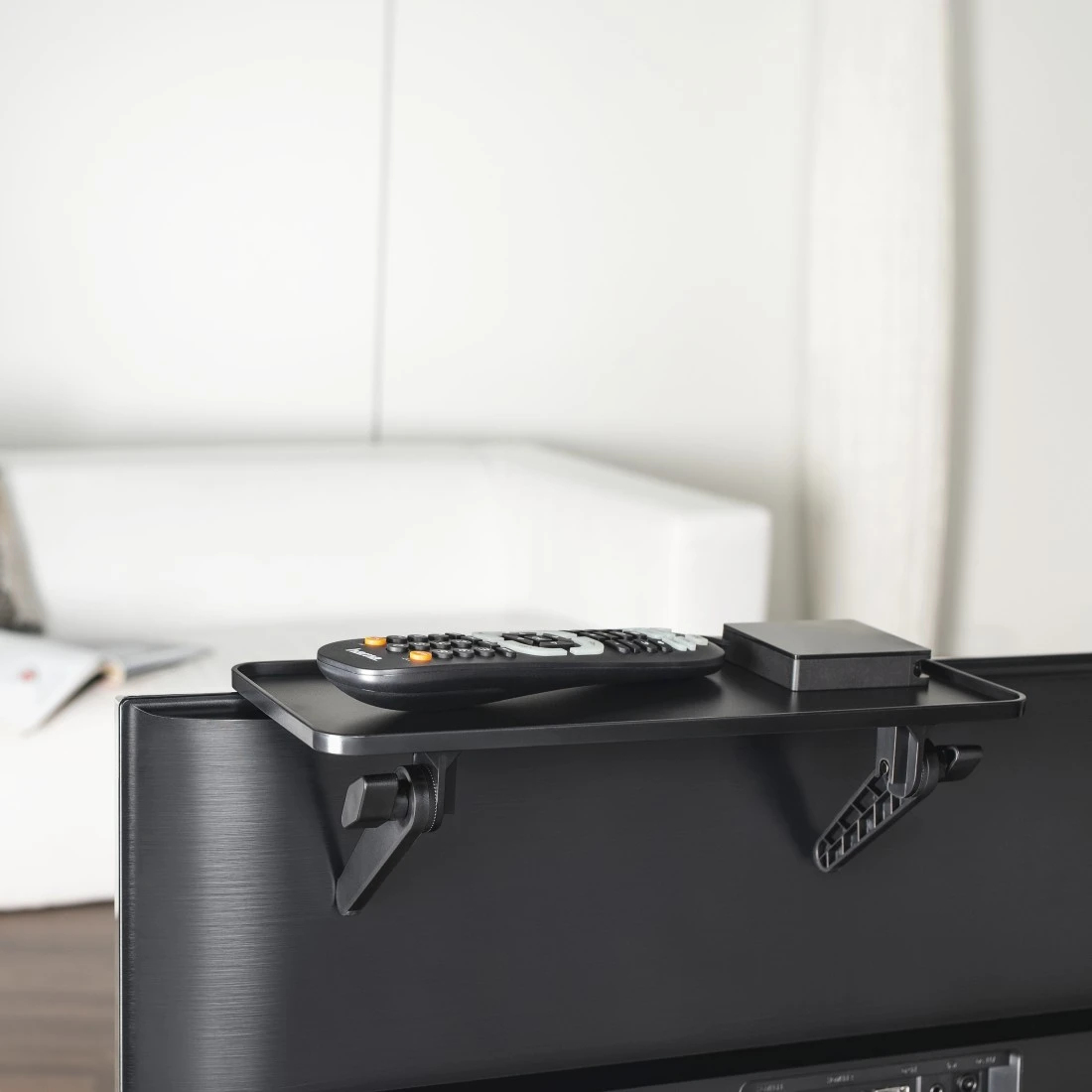 Telewizor z półką z pilotem i czarną skrzynką. Półka jest zamontowana na telewizorze.