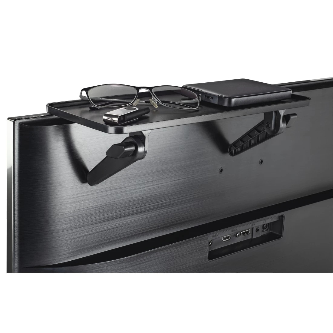 Telewizor z półką, na której leżą okulary, pendrive i czarne pudełko. Półka jest zamontowana z tyłu telewizora.