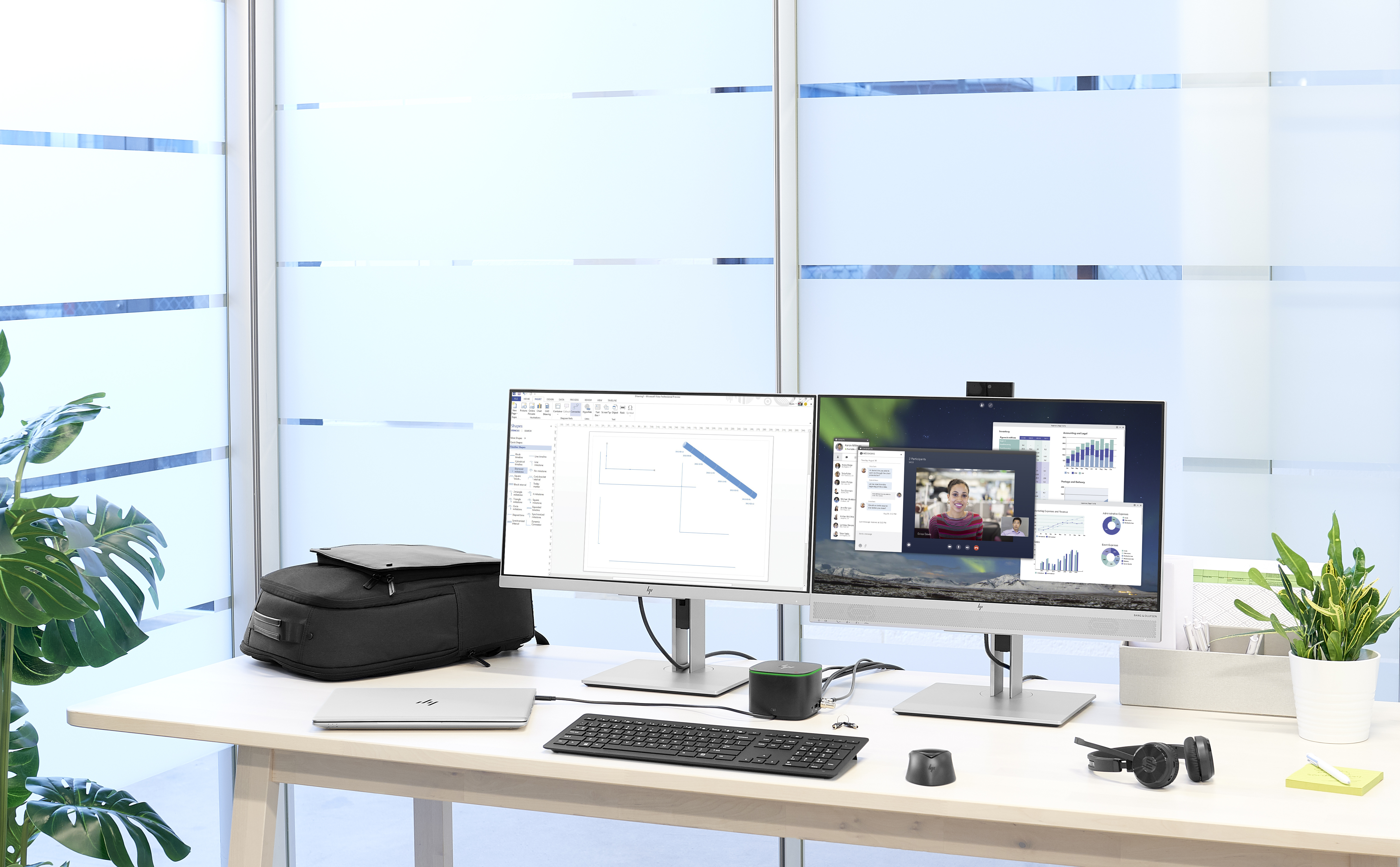 Configuración de oficina: dos monitores, portátil, teclado y planta en un escritorio blanco. Ventanas en el fondo.
