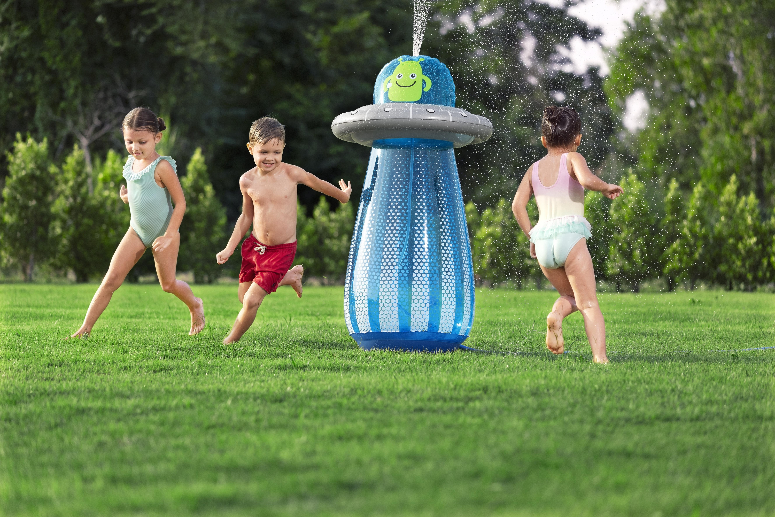 Troje dzieci biega przez zraszacze na zielonym trawniku. Pośrodku znajduje się nadmuchiwany zraszacz w kształcie kosmity.