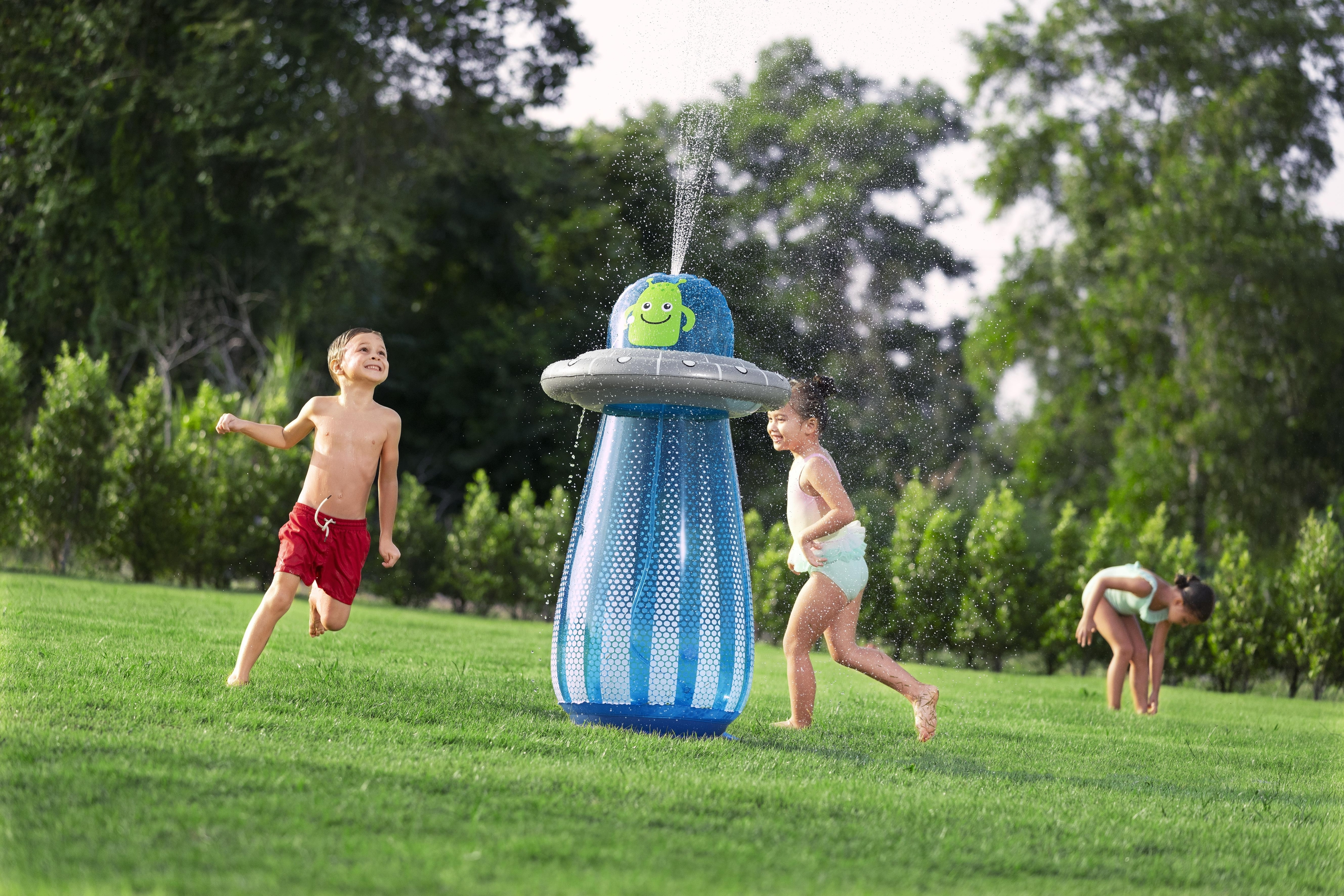 Trójka dzieci bawiących się na podwórku z zraszaczem wody w kształcie kosmity. Zielona trawa i drzewa w tle.