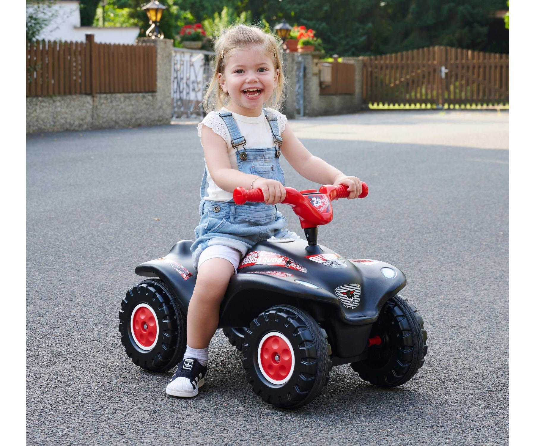Ein lächelndes Kind auf einem schwarzen Spielzeug-ATV mit roten Akzenten. Das Kind trägt eine Jeanslatzhose und sitzt auf einer gepflasterten Oberfläche.