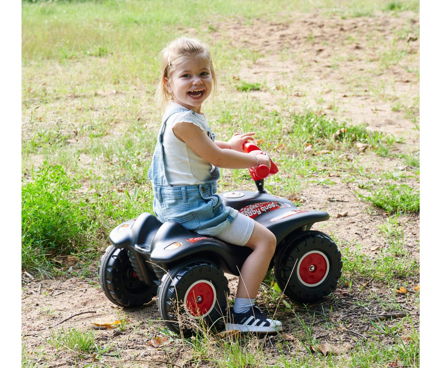 Ein lächelndes Kind sitzt auf einem schwarzen Spielzeug-ATV mit roten Rädern in einer grasbewachsenen Gegend.