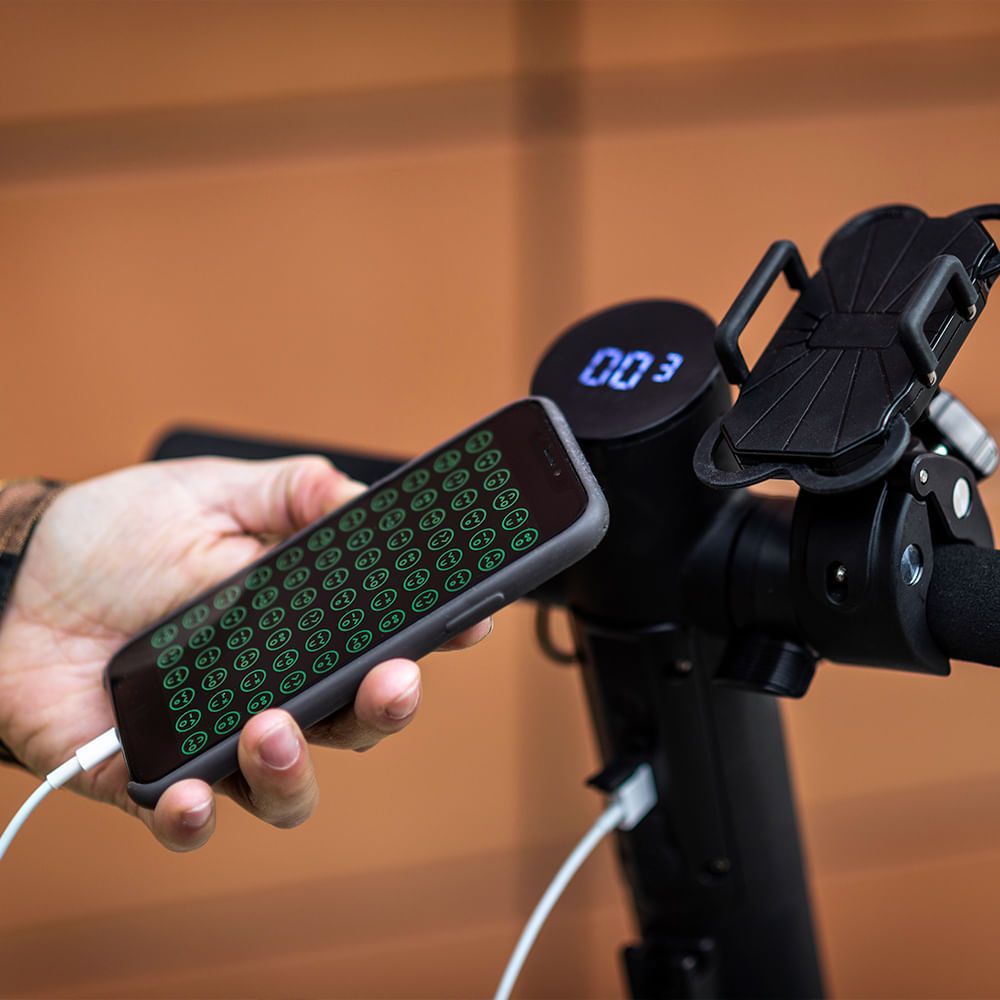 Una mano sostiene un teléfono cargando. La pantalla muestra iconos verdes. La pantalla de un patinete negro muestra '003'. Hay un soporte para teléfono.