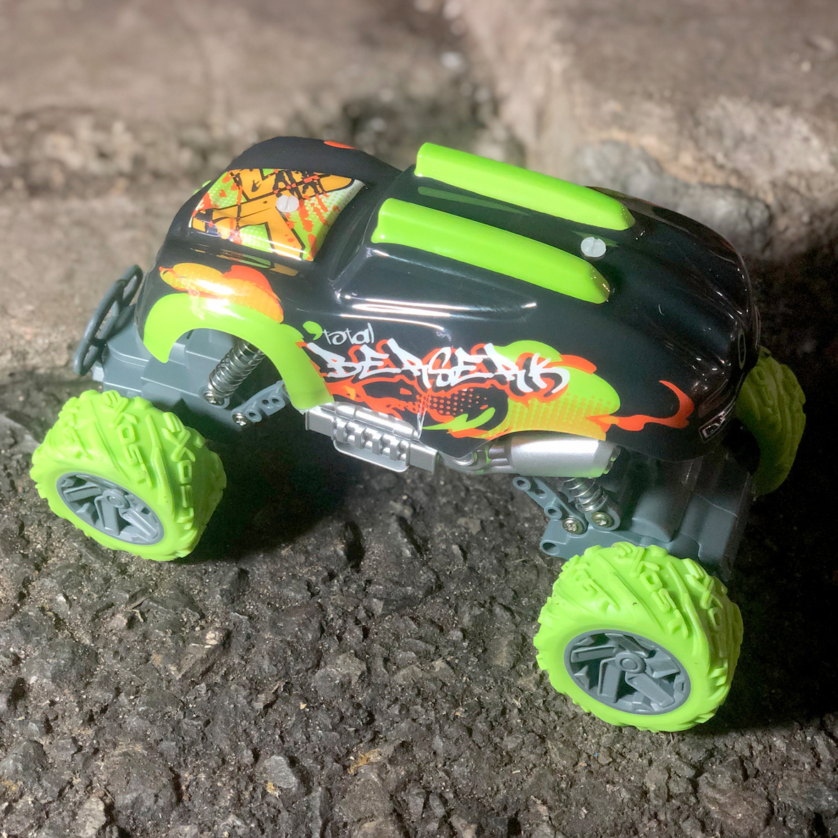 Un camión monstruo de juguete está sobre una superficie gris. Es negro con graffiti verde y naranja. Los neumáticos son verdes.