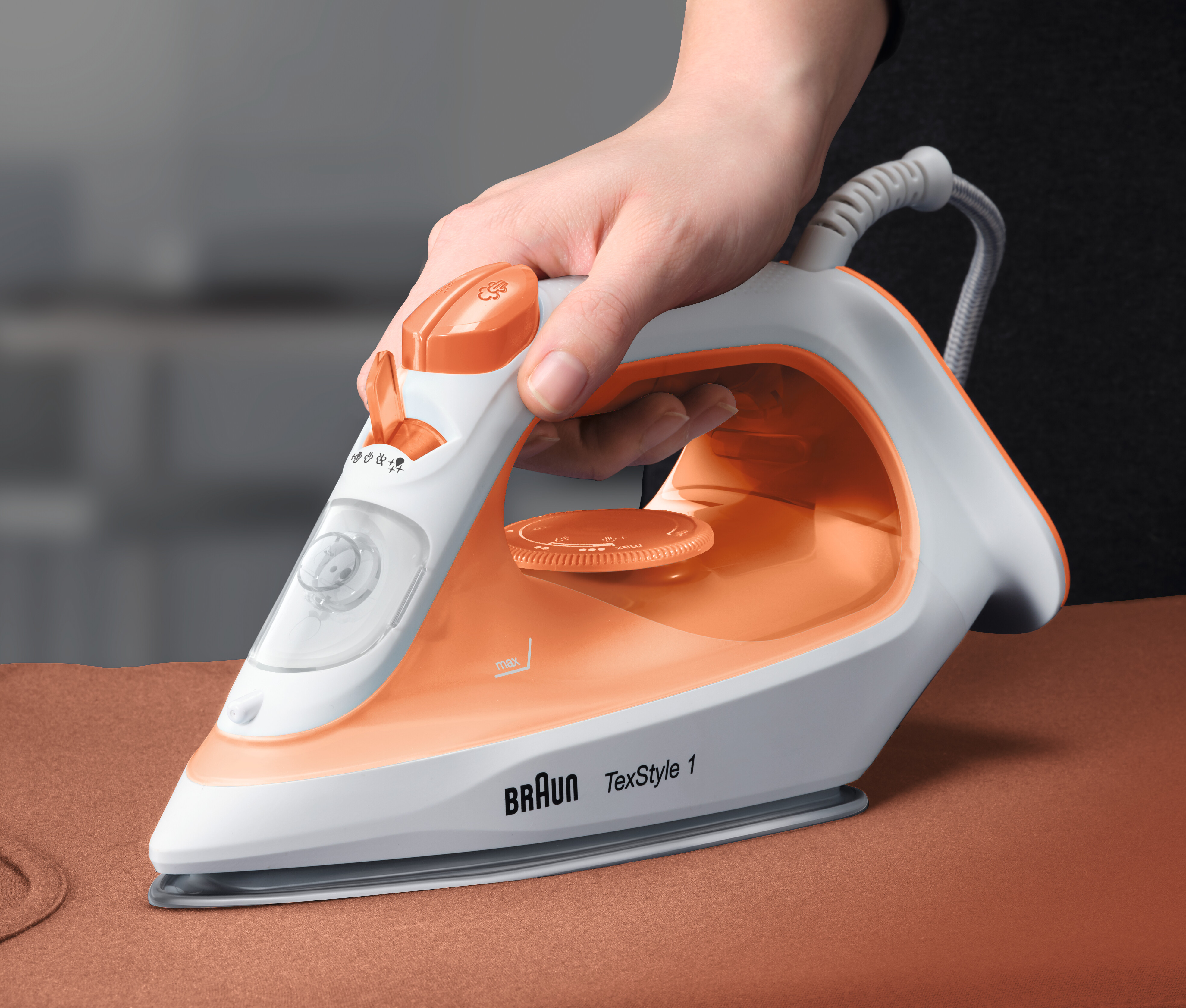 Ein weißes und orangefarbenes Braun TexStyle 1 Bügeleisen wird von einer Hand über braunem Stoff gehalten.