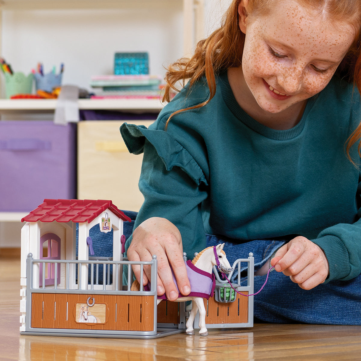 Dziewczyna bawi się zabawką-stajnią i białym koniem. Stajnia jest brązowo-biała z czerwonym dachem. Dziewczyna się uśmiecha.