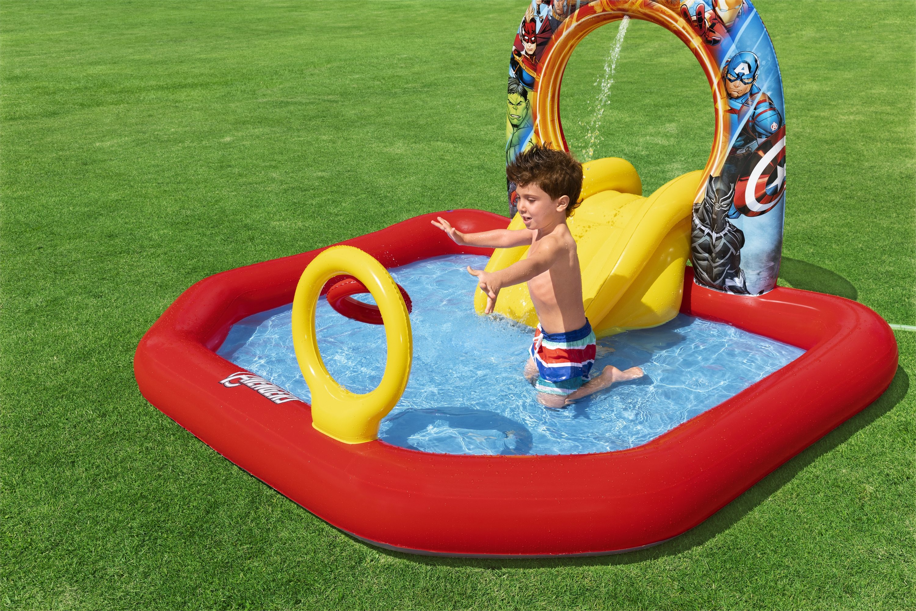 Dziecko bawi się w czerwonym i żółtym basenie Avengers, woda tryska z góry, na zielonej trawie.