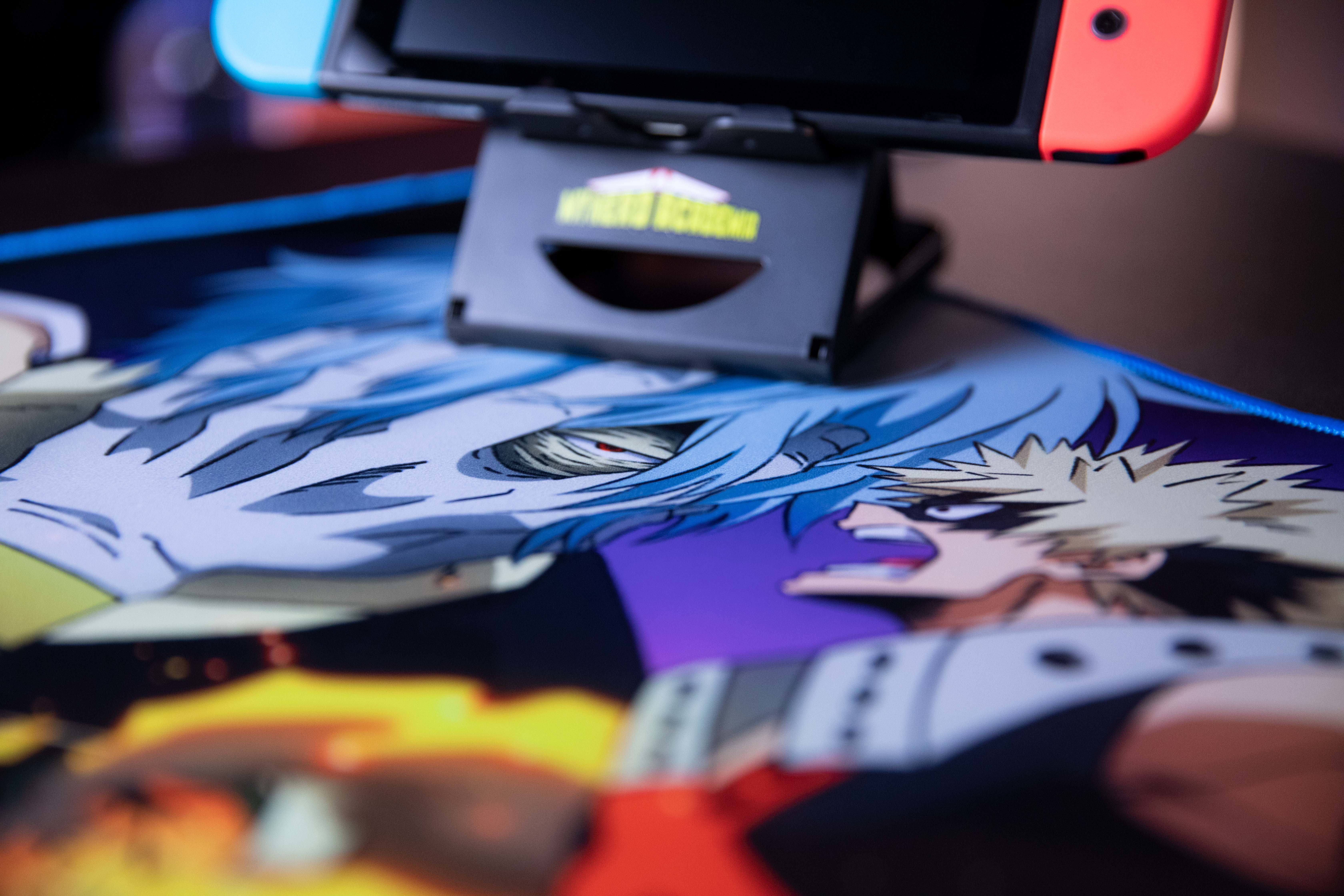 Konsola Switch na podstawce. Poniżej podkładka pod mysz z postaciami z anime. Kolory: czarny, niebieski, pomarańczowy i fioletowy.