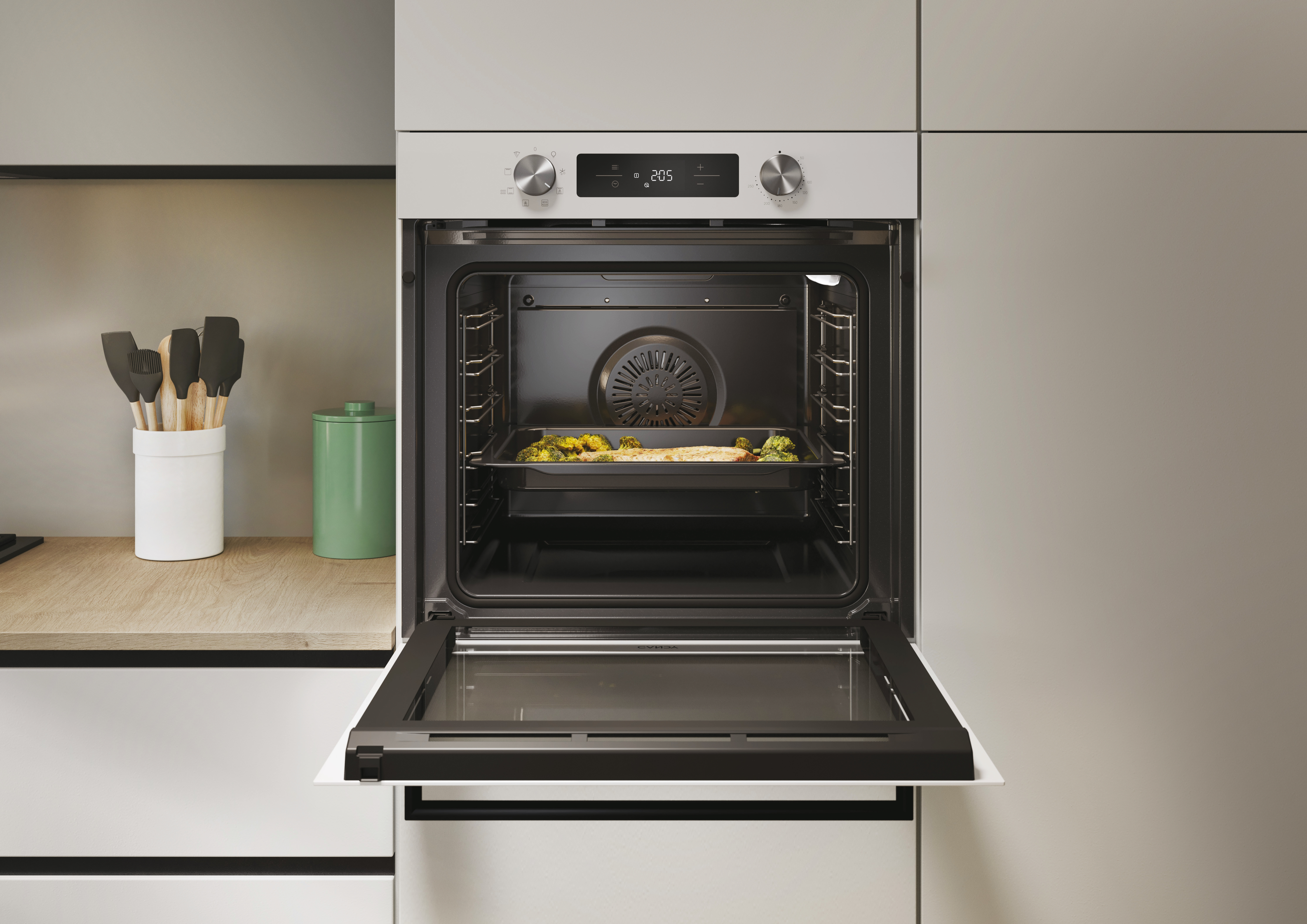 Otwarty piekarnik z jedzeniem na tacy. Przybory kuchenne na blacie. Piekarnik biały z czarnymi drzwiami.