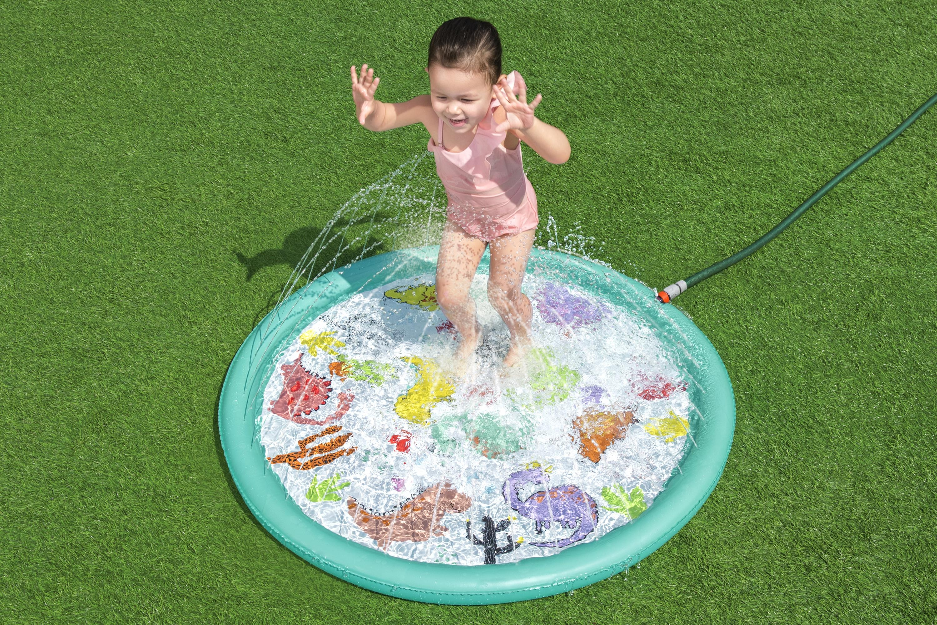 Una bambina in costume da bagno rosa salta in una piscina per bambini su erba verde. Il cuscinetto ha disegni di dinosauri.