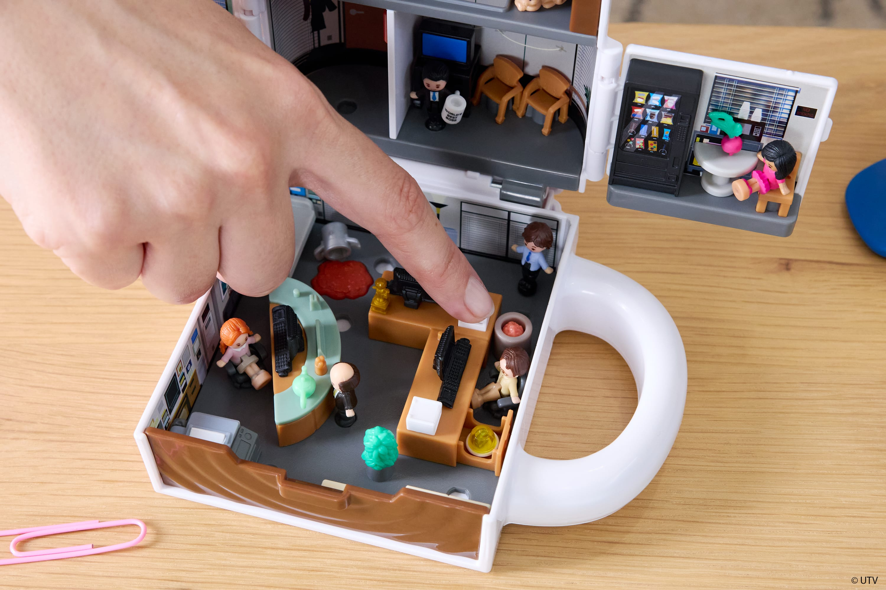Ręka wskazuje na miniaturowy zestaw biurowy. W środku znajdują się małe figurki, meble i automat. Zestaw stoi na drewnianym stole.