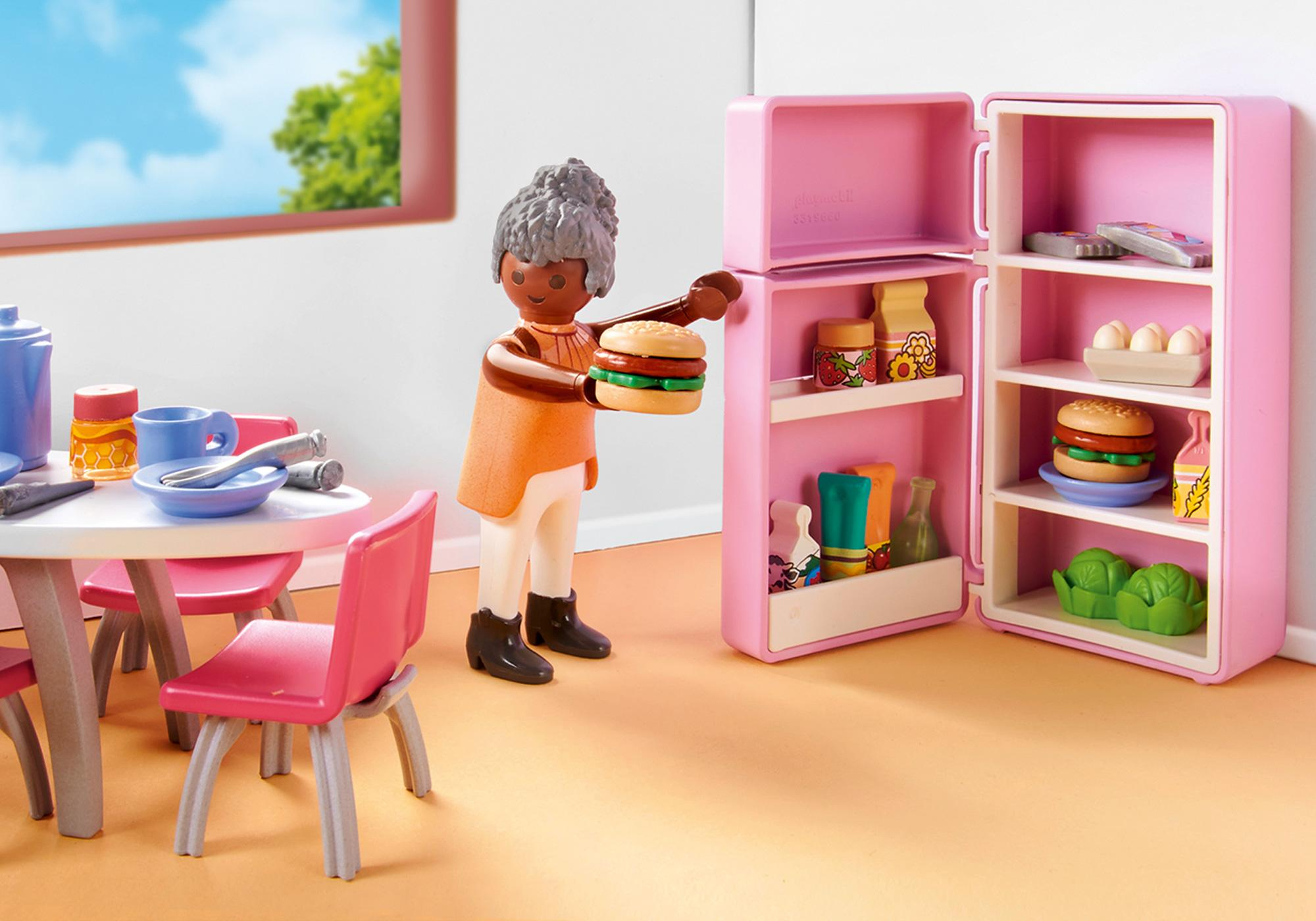 Eine Spielfigur hält einen Burger in der Nähe eines offenen, pinkfarbenen Kühlschranks. Ein Tisch mit Stühlen und Essen befindet sich im Hintergrund. Links ist ein Fenster.