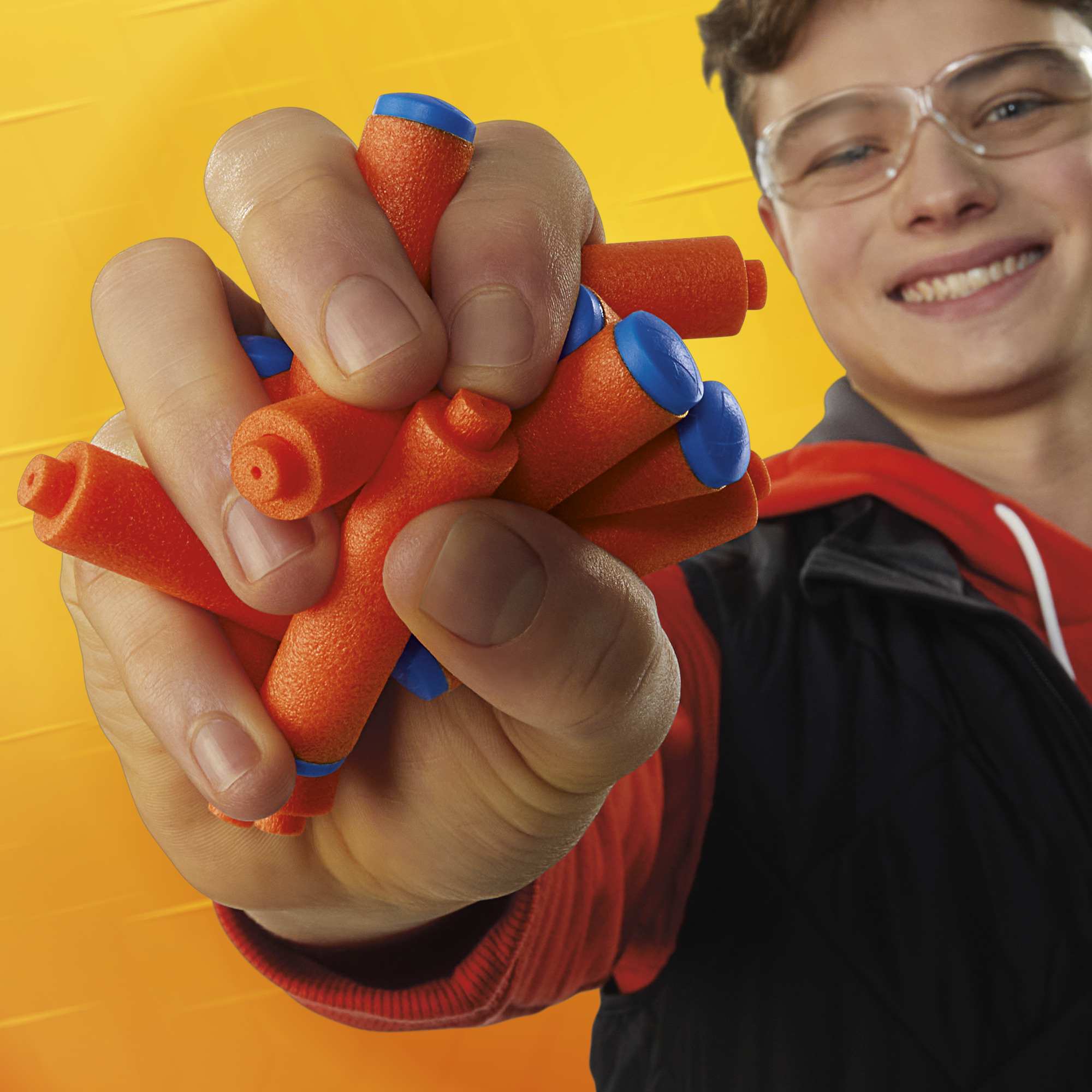 Una mano sostiene dardos de espuma naranja y azul. Un niño sonriente con gafas está en el fondo.
