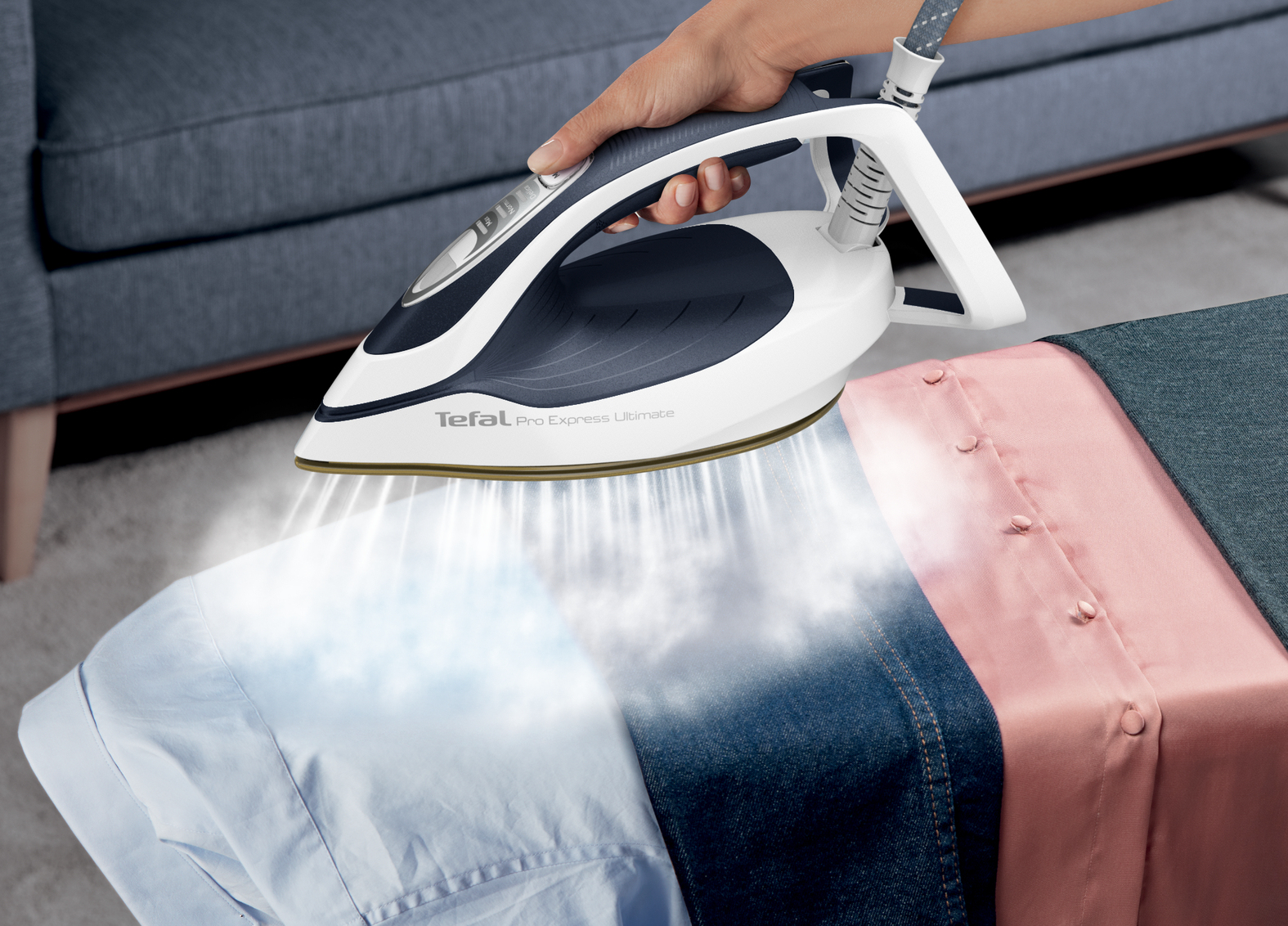 Eine Hand bügelt Kleidung mit einem Tefal-Bügeleisen. Dampf steigt vom Bügeleisen auf ein hellblaues Hemd, Jeans und ein rosa Hemd.