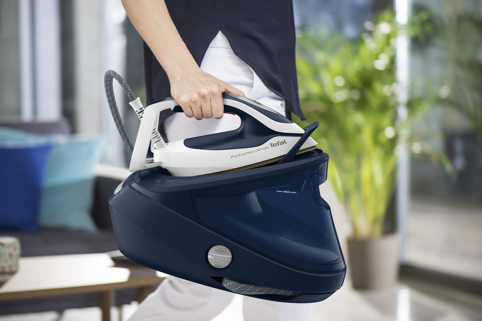 Eine Person hält ein Tefal-Bügeleisen. Das Bügeleisen ist weiß und marineblau und ruht auf seiner marineblauen Basis. Der Hintergrund zeigt einen Raum.