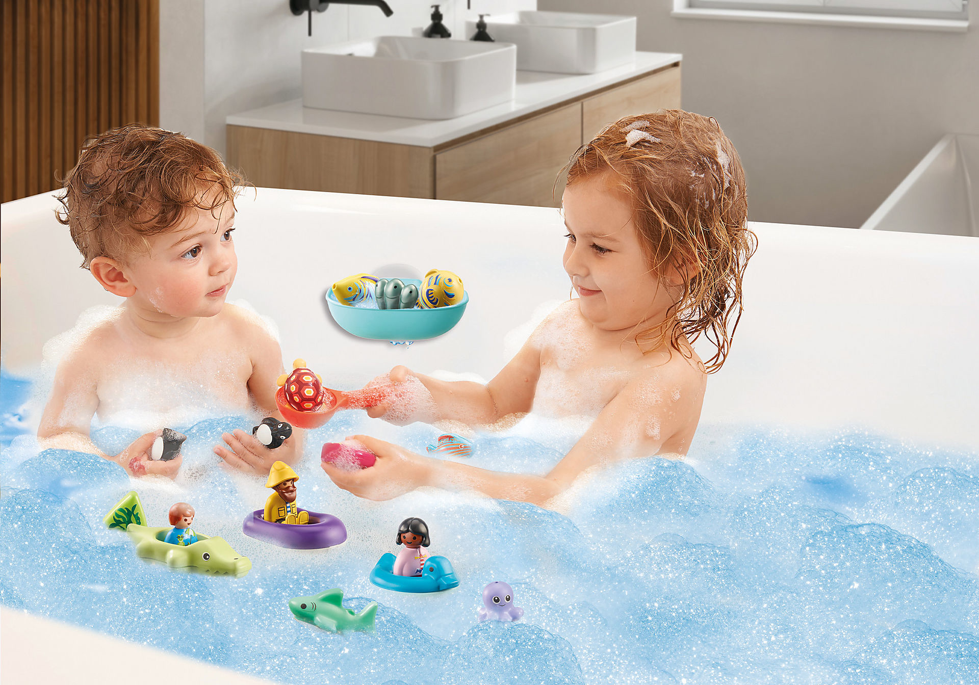 Due bambini in una vasca da bagno con bolle e giocattoli. Ci sono barche giocattolo, animali e un vassoio blu con giocattoli di pesci. Lo sfondo mostra un bagno moderno.