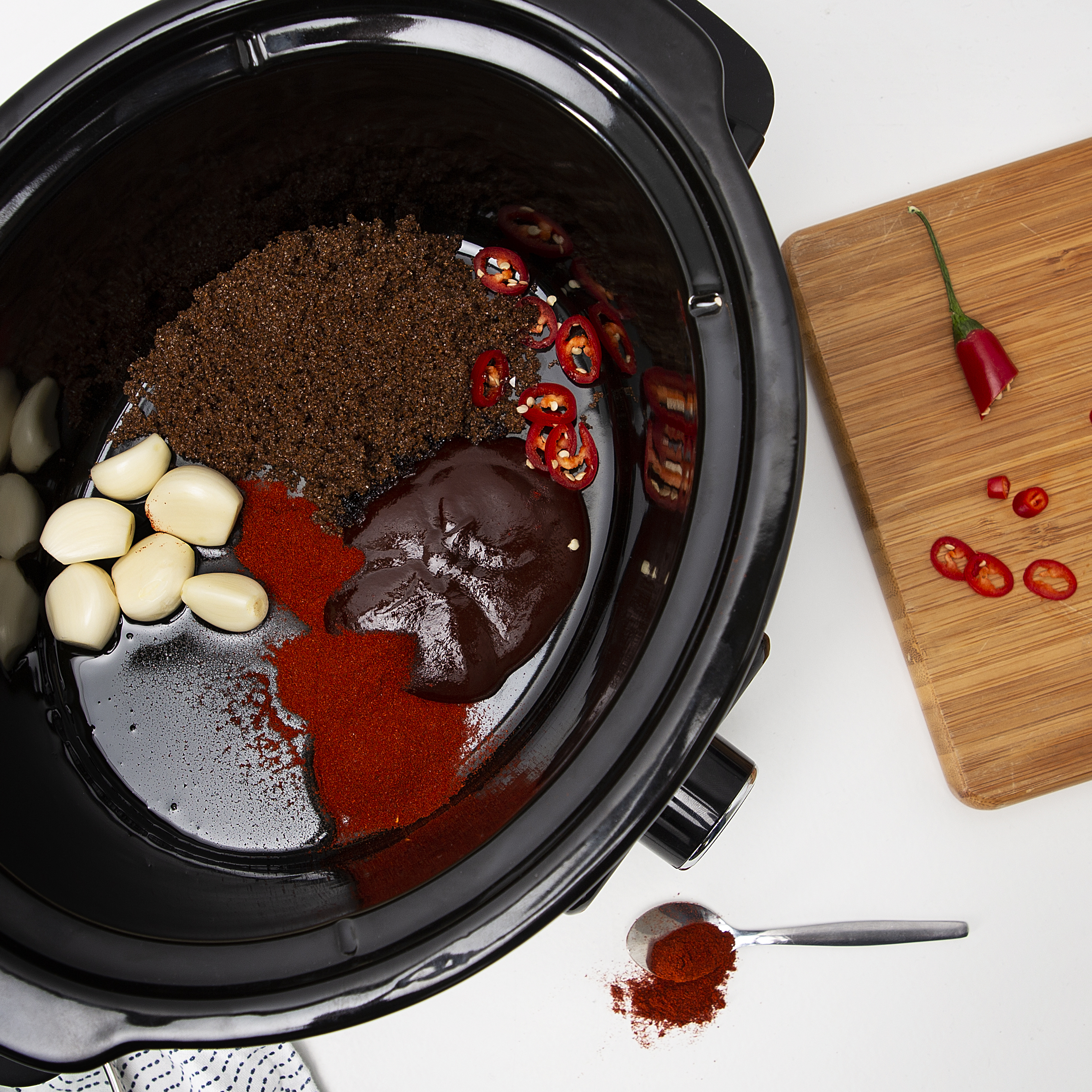 Ein Schongarer enthält Zutaten: Knoblauch, Gewürze, geschnittene rote Paprika und ein Holzschneidebrett mit Pfeffer.