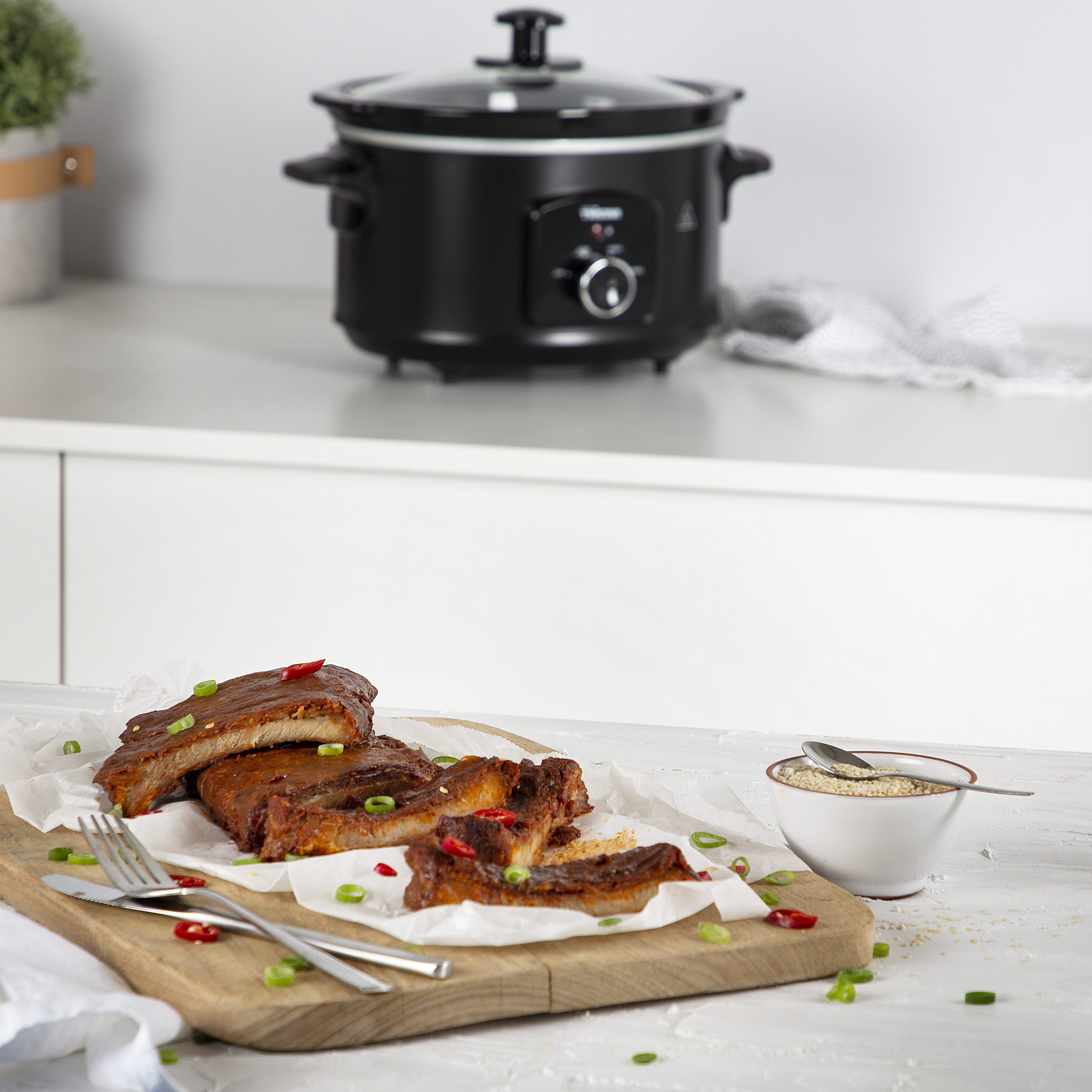 Gekochte Rippchen auf einem Holzbrett mit Chili und Frühlingszwiebeln, ein Slow Cooker im Hintergrund.