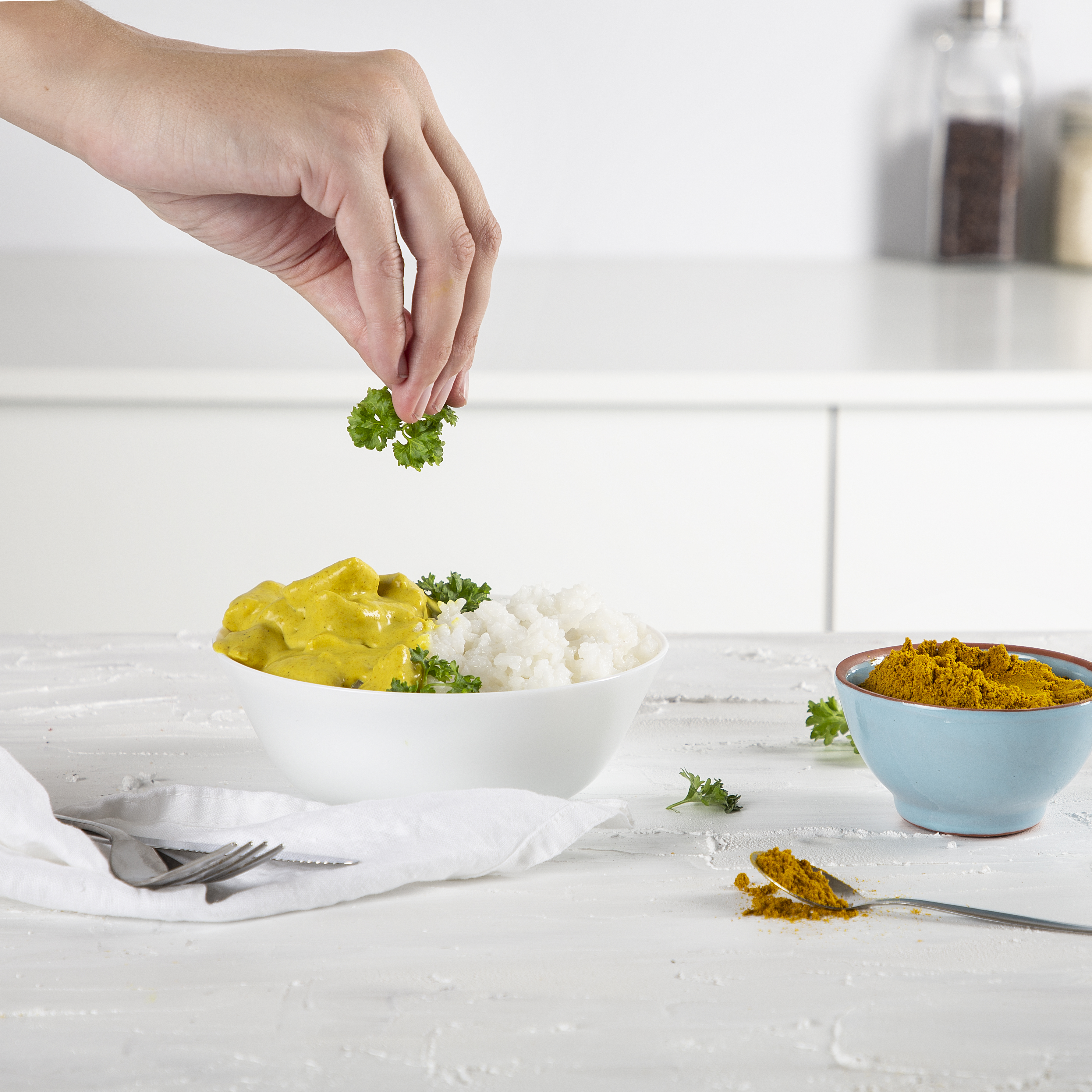 Eine Hand streut Petersilie über Curry und Reis in einer Schüssel. Eine Schüssel mit Currypulver steht daneben.