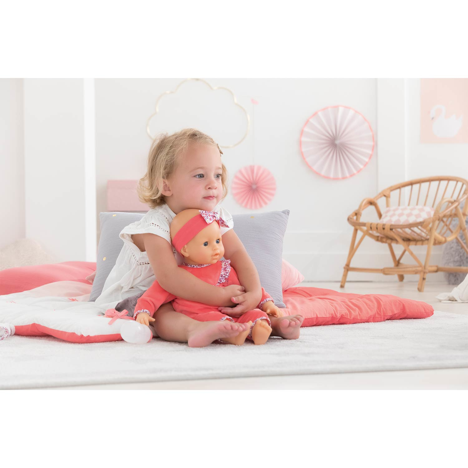 Ein junges Mädchen umarmt eine Puppe, beide in passenden korallenfarbenen Outfits. Sie sitzen auf einem weißen Teppich mit korallenfarbenen Kissen.