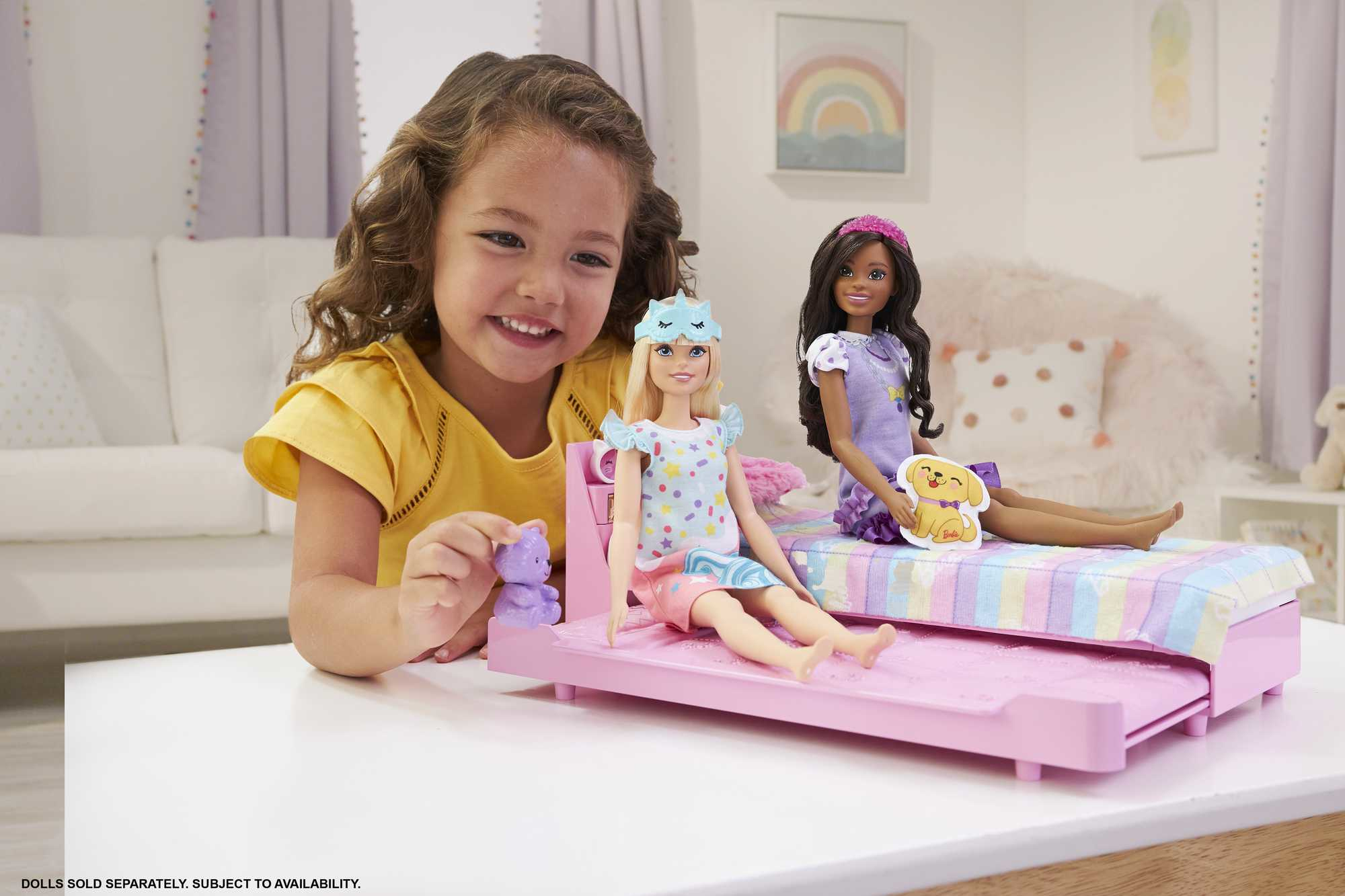 Una niña sonríe, sosteniendo un juguete. Dos muñecas sentadas en una cama rosa. Una tiene antifaz azul y la otra una diadema.