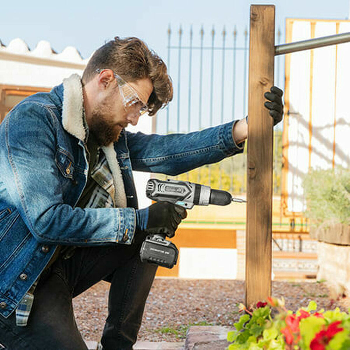 Un hombre con una chaqueta vaquera usa un taladro en un poste de madera. Usa gafas de seguridad y guantes. El fondo tiene plantas y una valla.